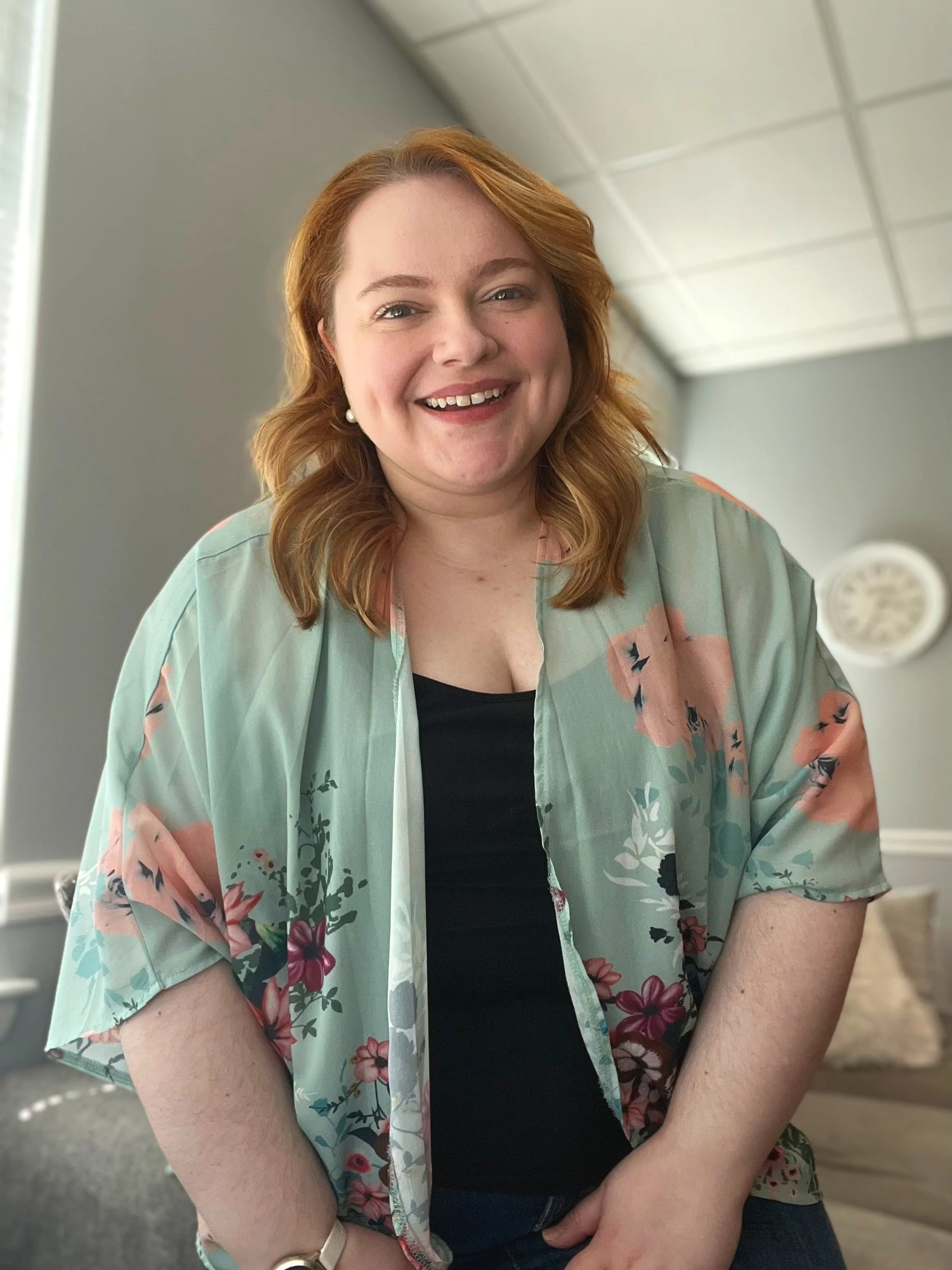 A woman with red hair smiling, wearing a floral cardigan over a black top, standing indoors with a clock on the wall in the background.