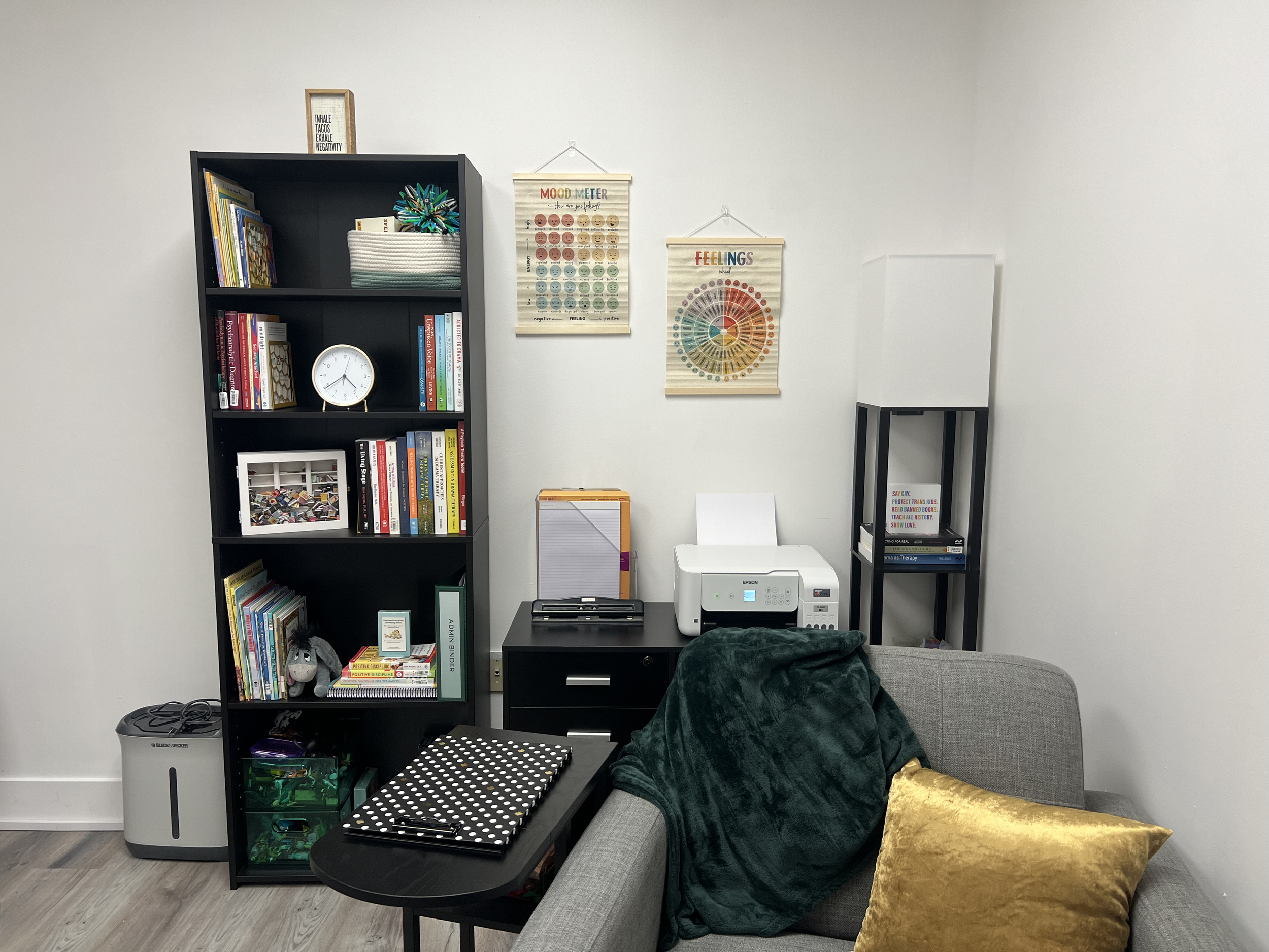 Office corner with black bookshelf filled with books and decorative items, white clock, and framed photos. To the right, a small black desk with a polka-dotted notebook, a printer, and a whiteboard. Gray sofa with green blanket and yellow pillow. Two posters on the wall about mood meter and feelings.