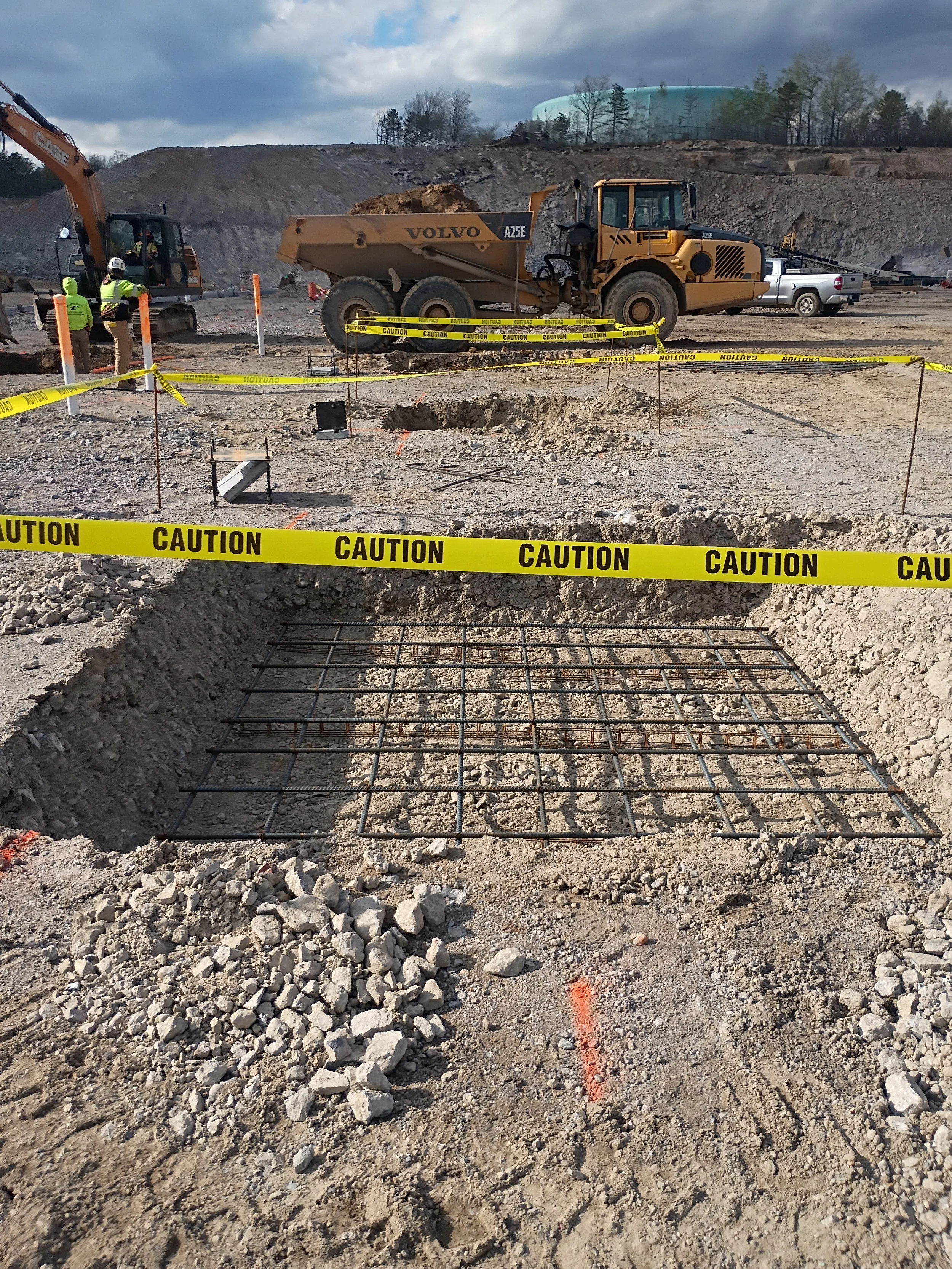 Construction site with yellow caution tape, rebar, and excavator in the background. Uneven ground and rocks in the foreground.