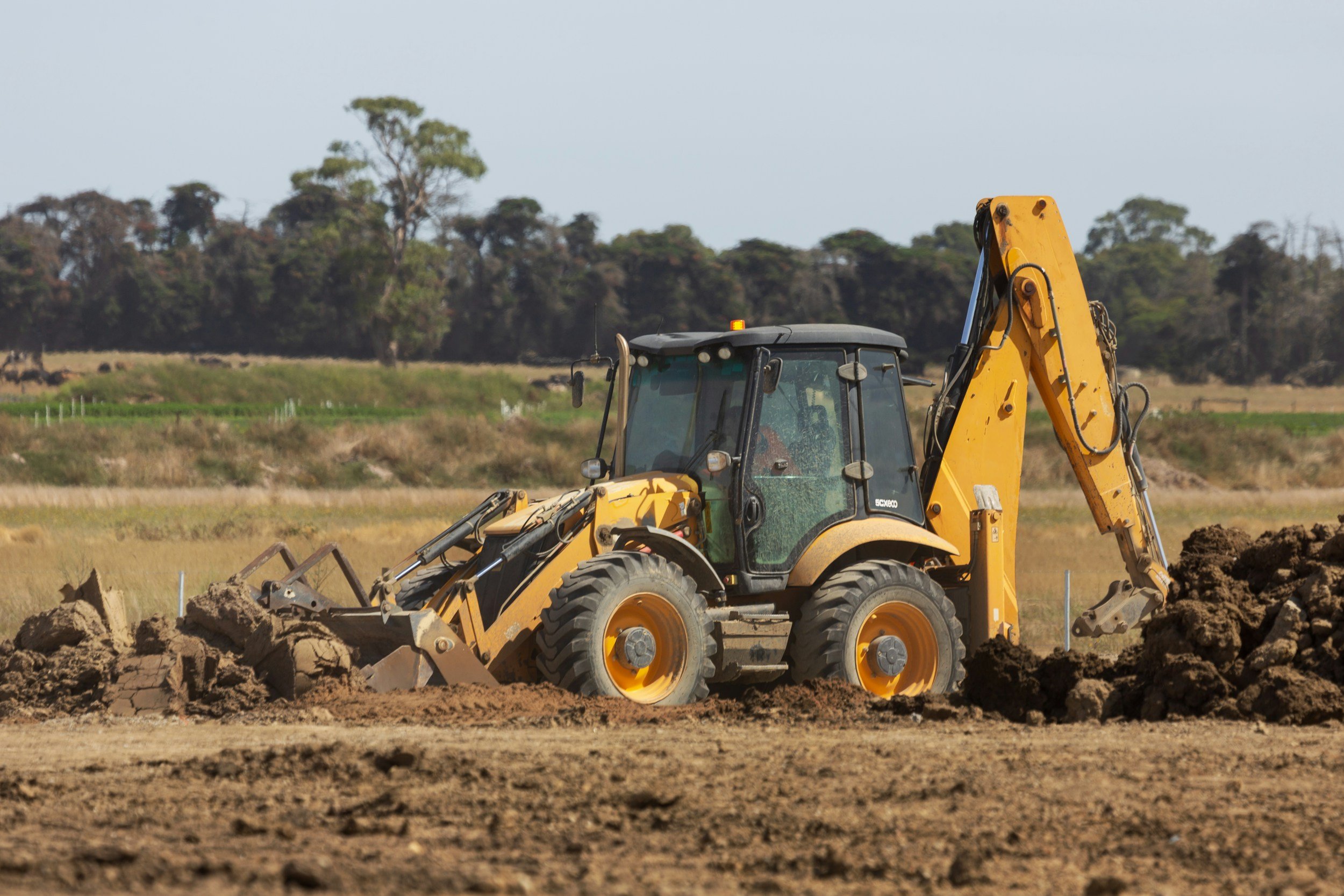 Yellow backhoe excavator digging in dirt on a ranch, with trees and open fields in the background.