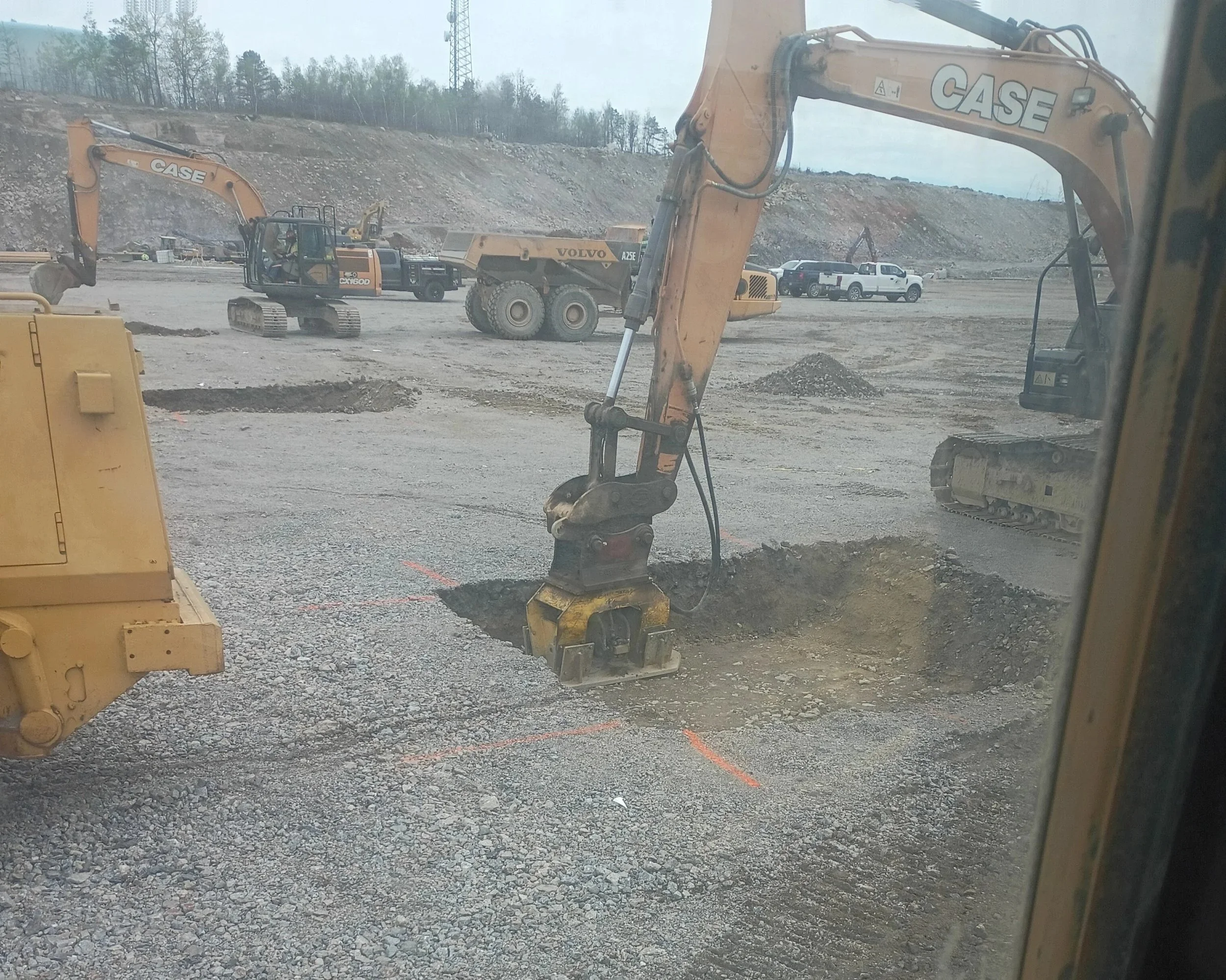 Construction site with heavy machinery including two excavators, one with the brand name CASE and another with the brand name Volvo, and several trucks in the background, dirt mounds, and trees on a cloudy day.