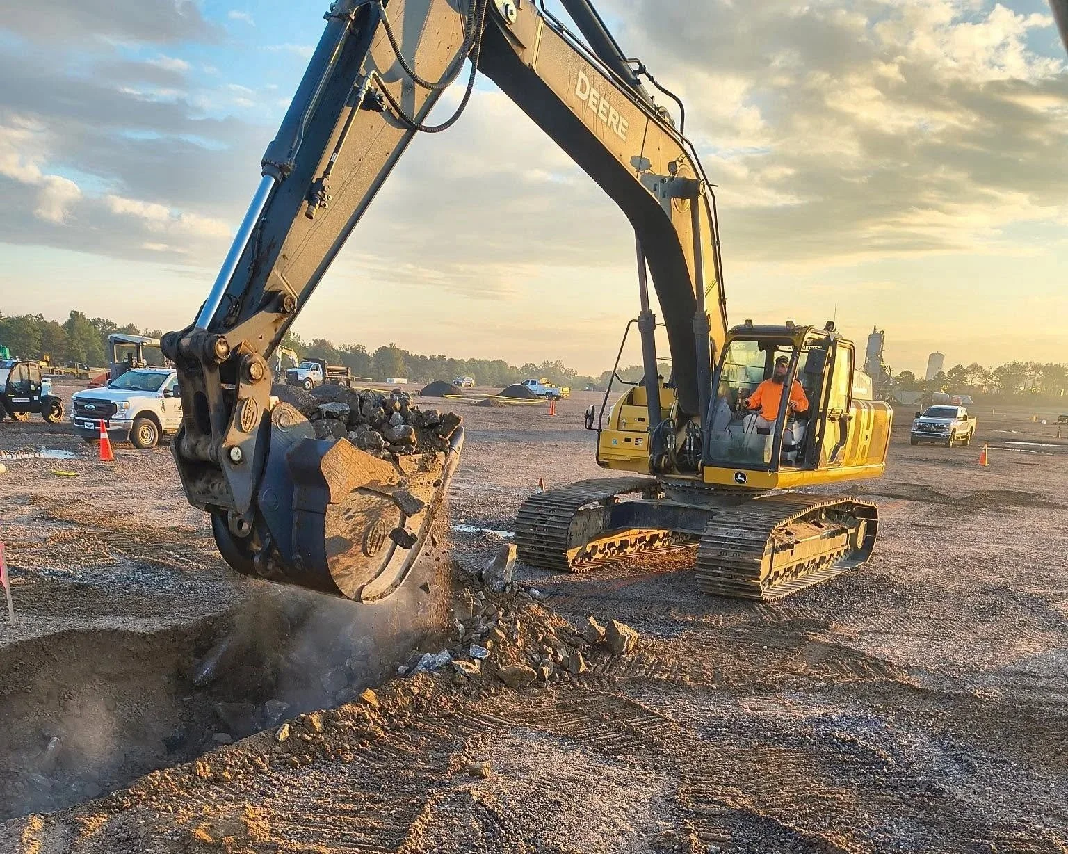 A yellow excavator is digging into the ground at a construction site during sunset, with several cars and trucks parked in the background.