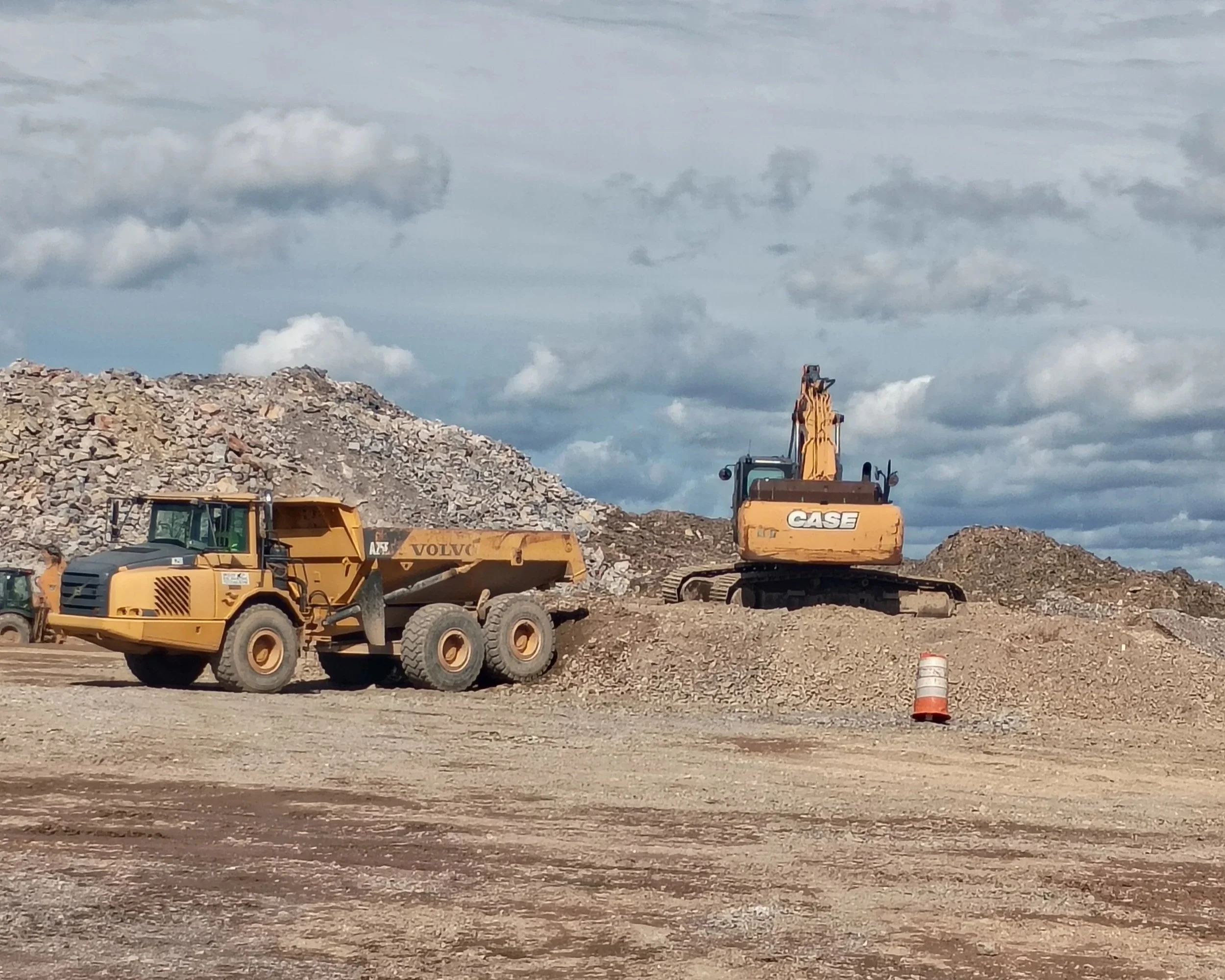 Construction site with large pile of rocks, a yellow front loader, and a yellow excavator under a partly cloudy sky.
