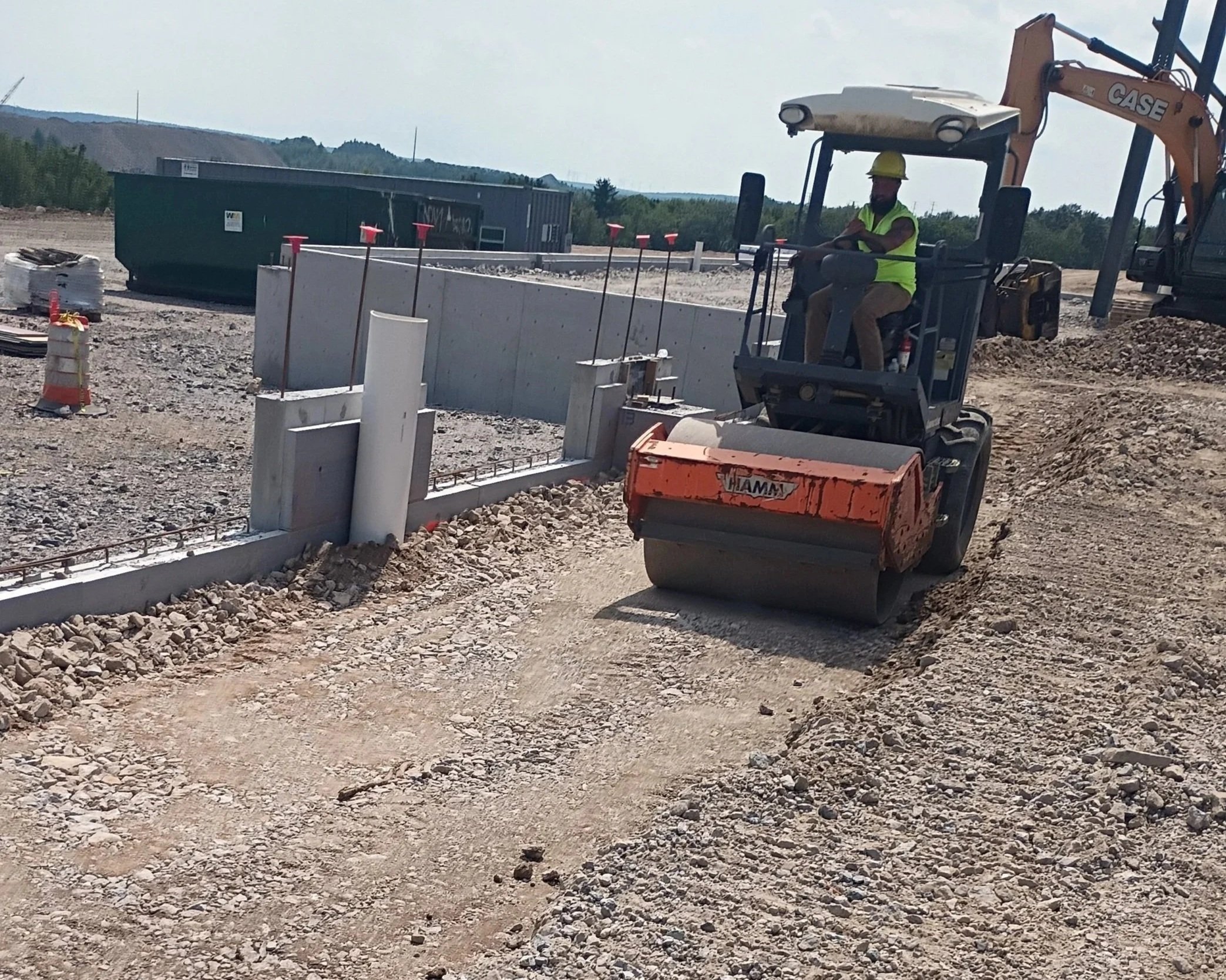 Construction worker operating a mini excavator on a building site with piles of gravel and construction materials around.