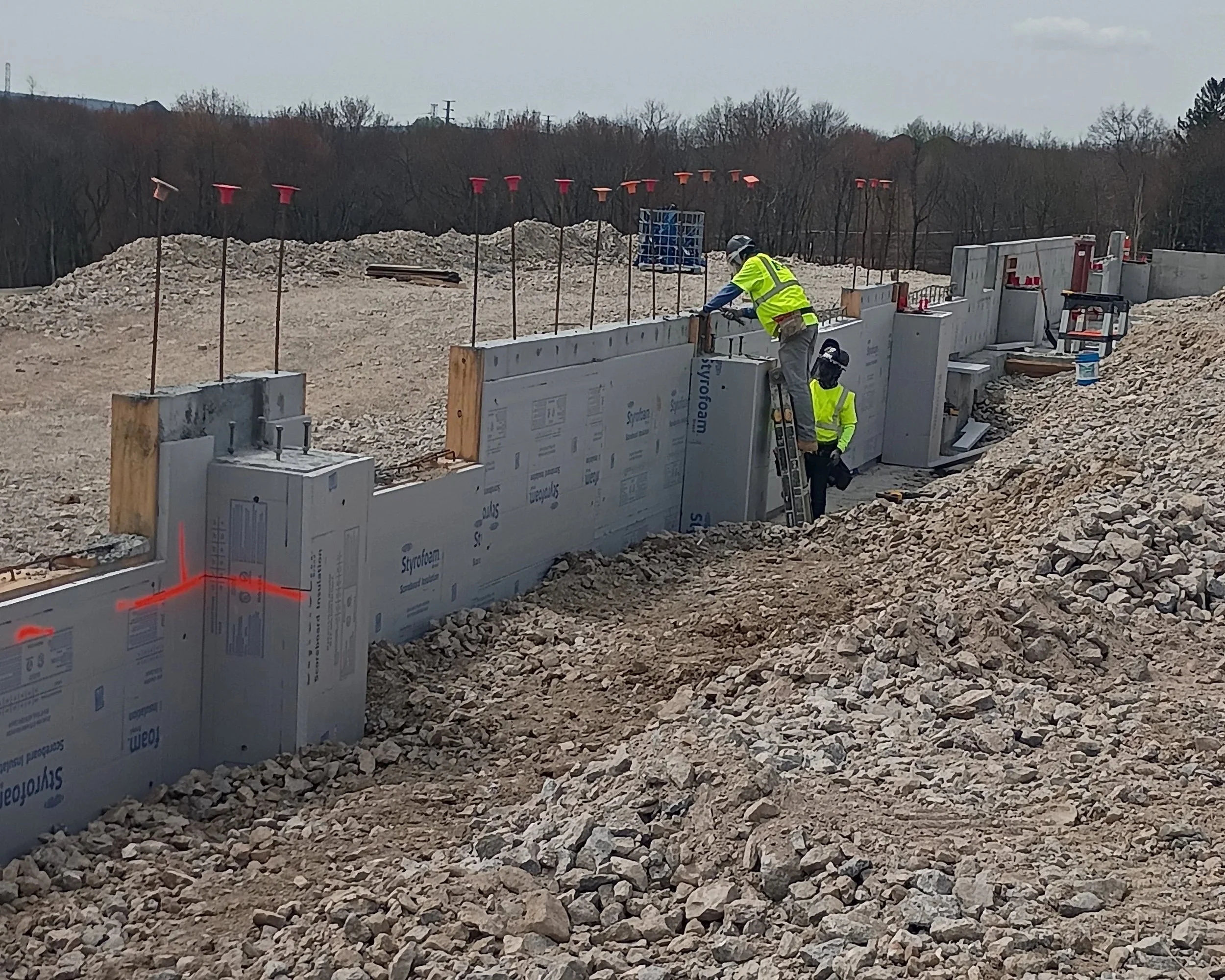 Construction workers in high-visibility vests and helmets working on a building foundation, with concrete blocks and rocky ground, and orange markers in the background.