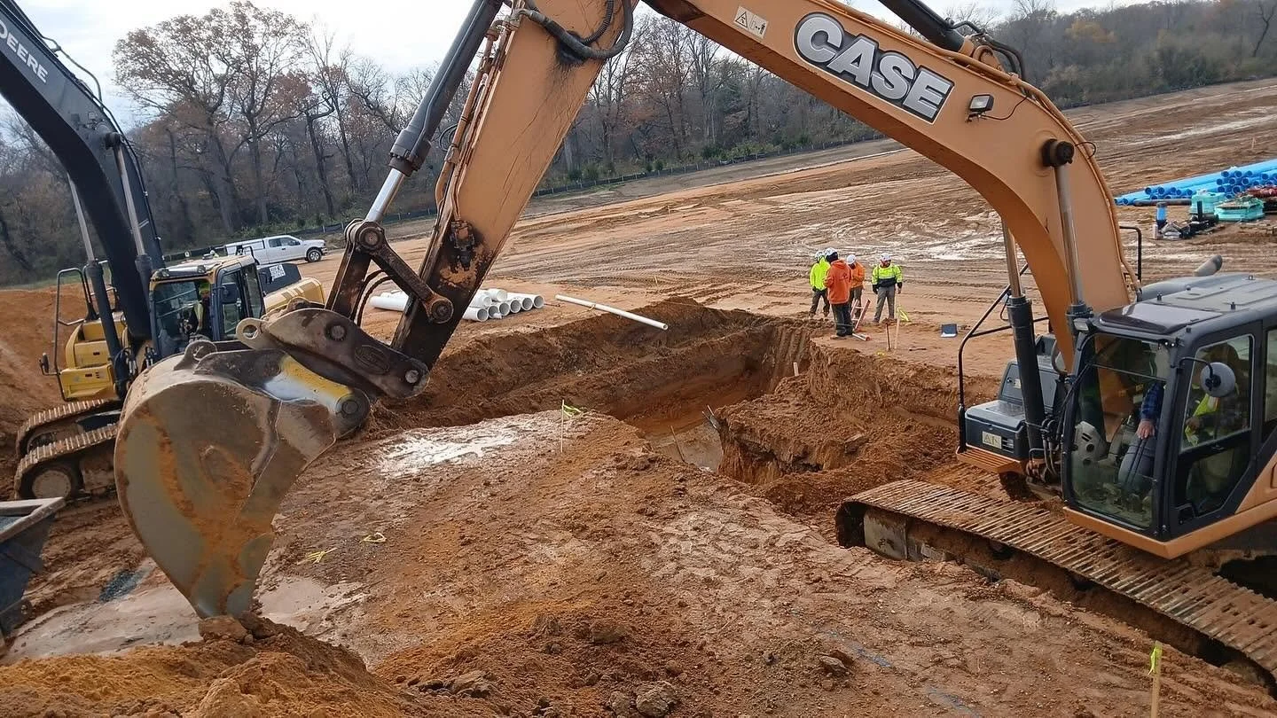 Construction site with excavators and workers digging a trench in dirt, with trees and vehicles in the background.