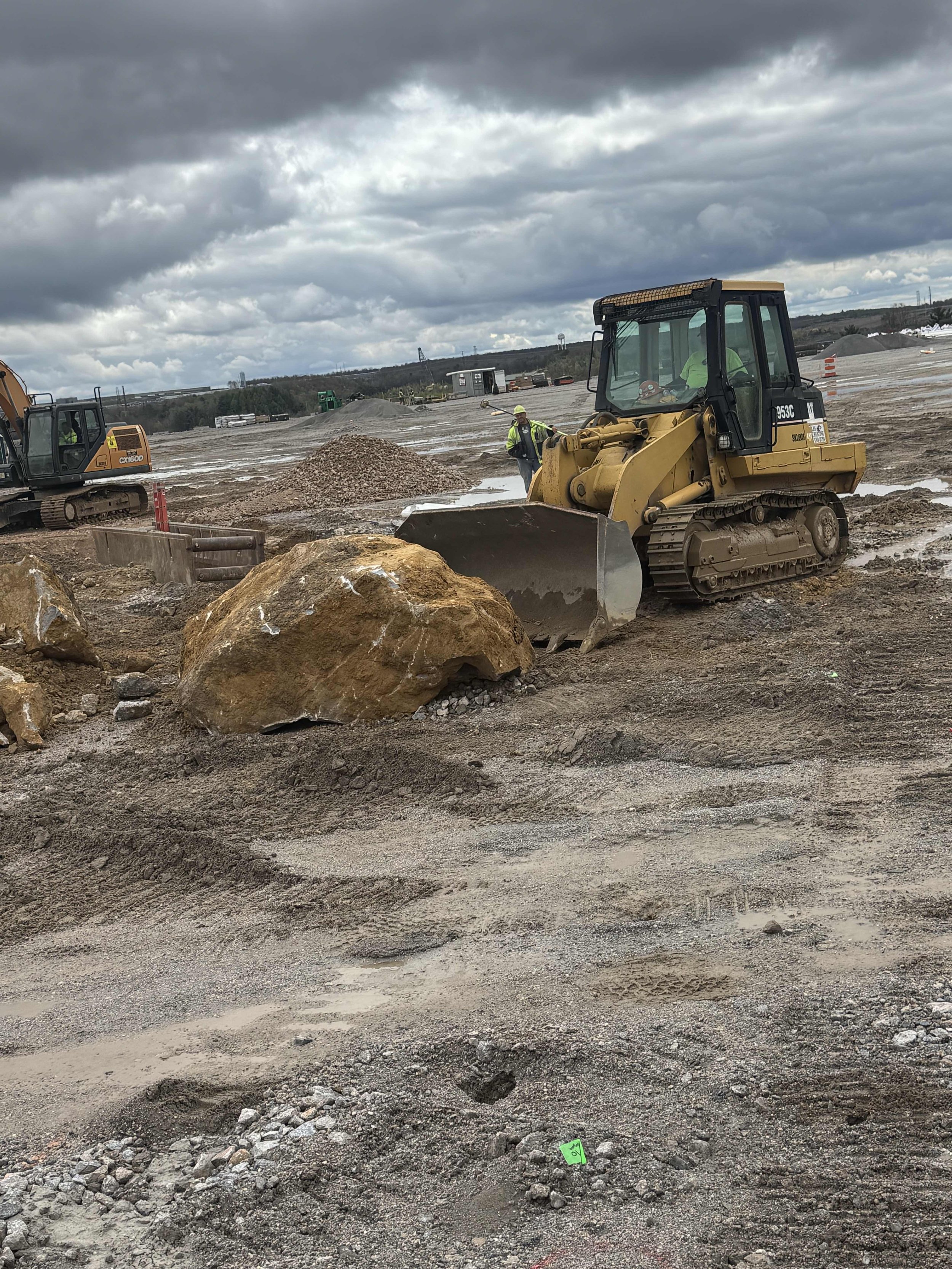 Construction site with heavy machinery, workers in safety gear, large rocks, and cloudy sky.