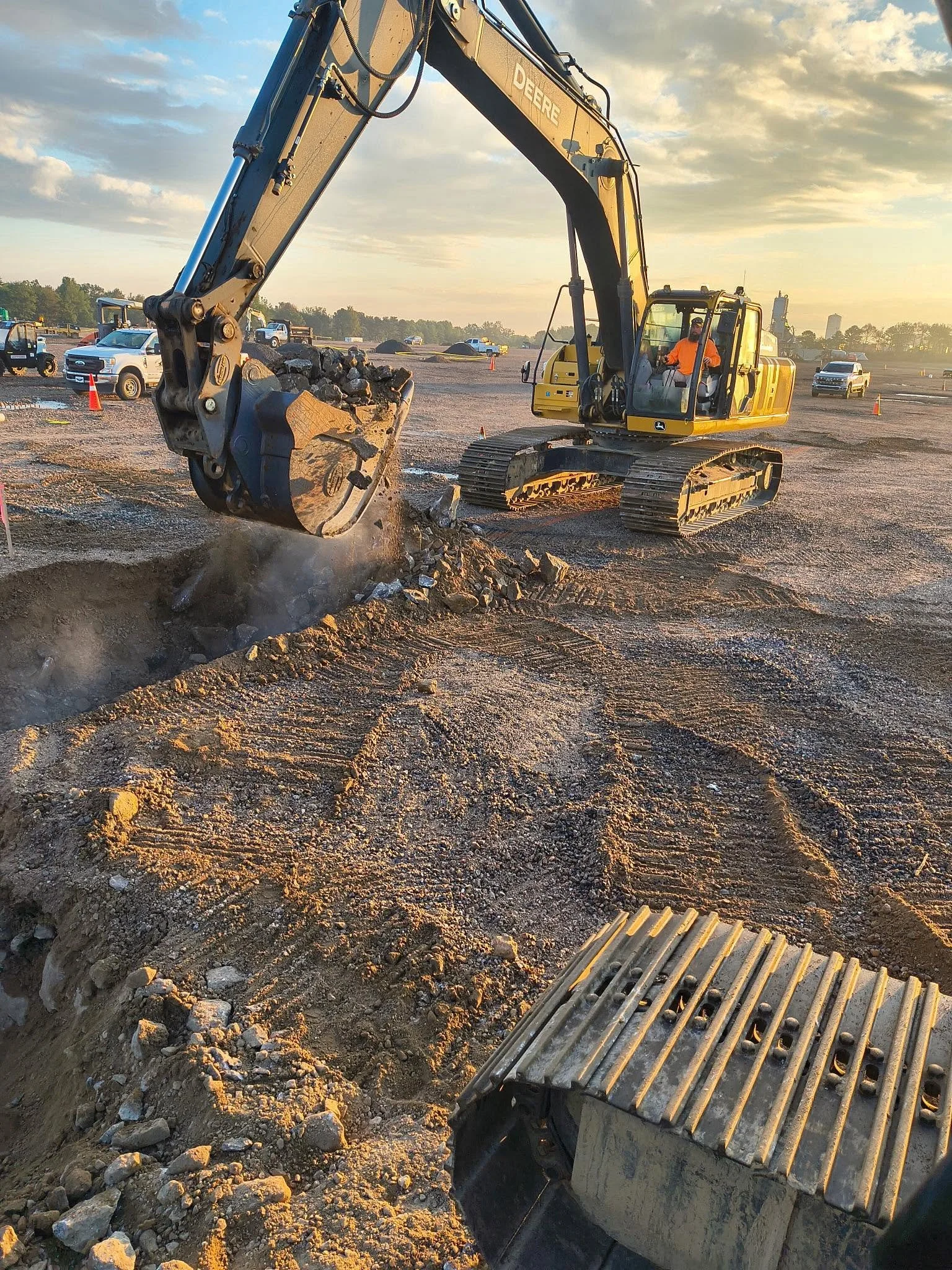 A construction excavator lifting rocks at a construction site during sunset with several parked vehicles and a cloudy sky in the background.