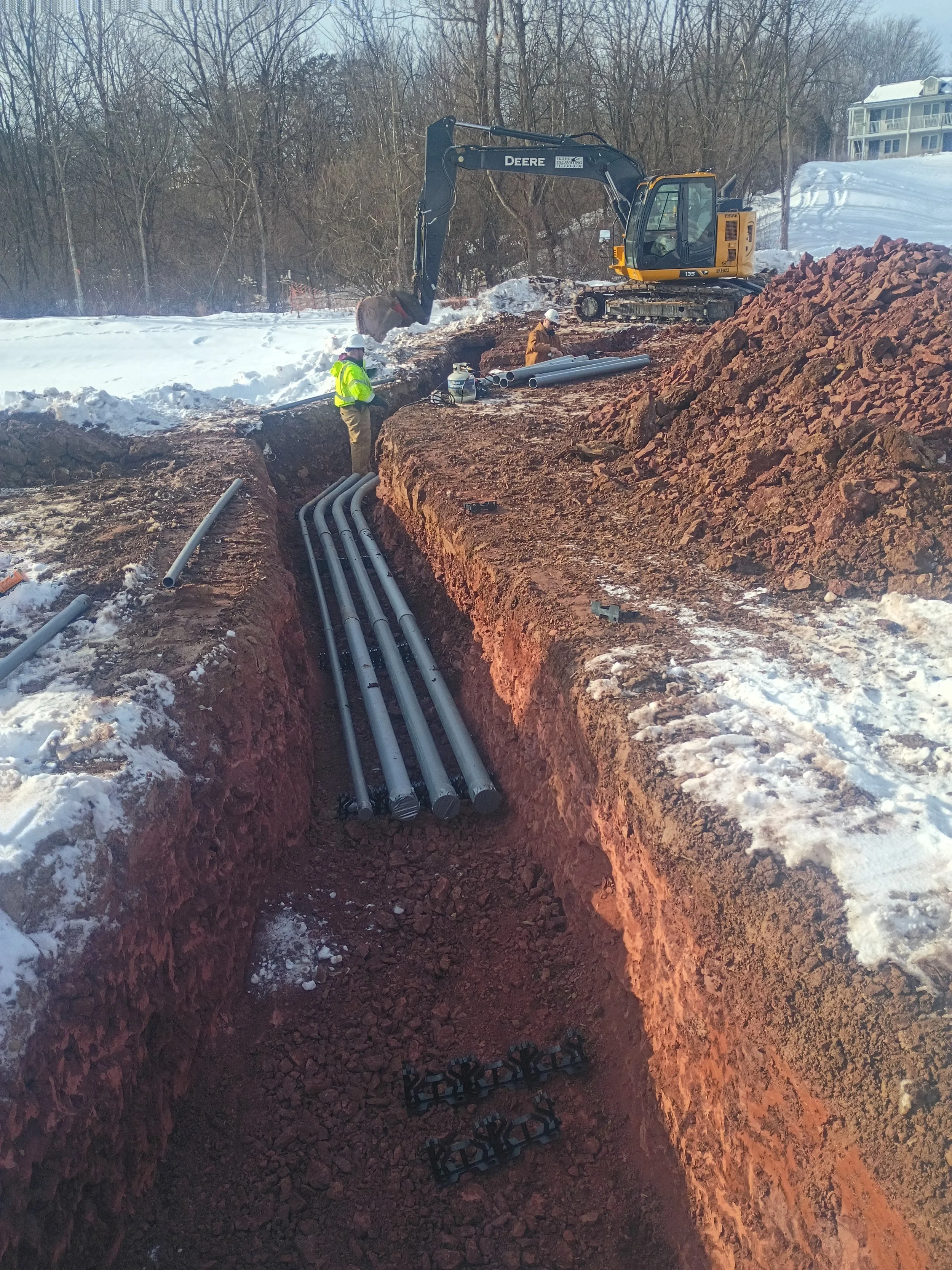 Construction workers installing underground pipes in a snowy area, with a John Deere excavator and a residential building in the background.