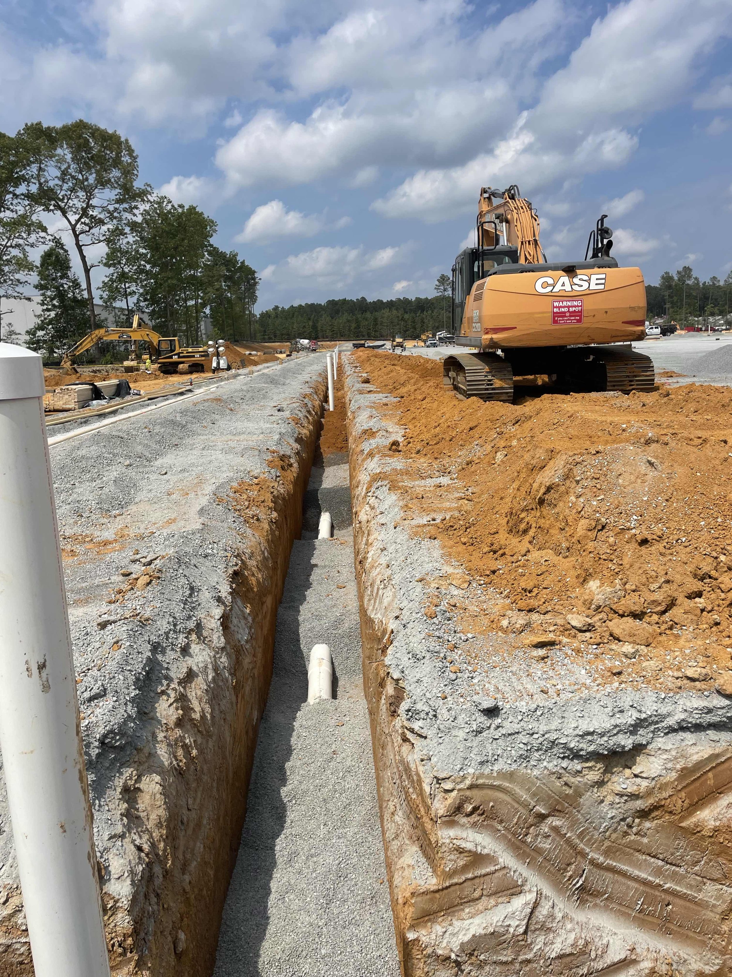 Construction site with a large orange excavator, a trench, gravel, and construction equipment under a partly cloudy sky.