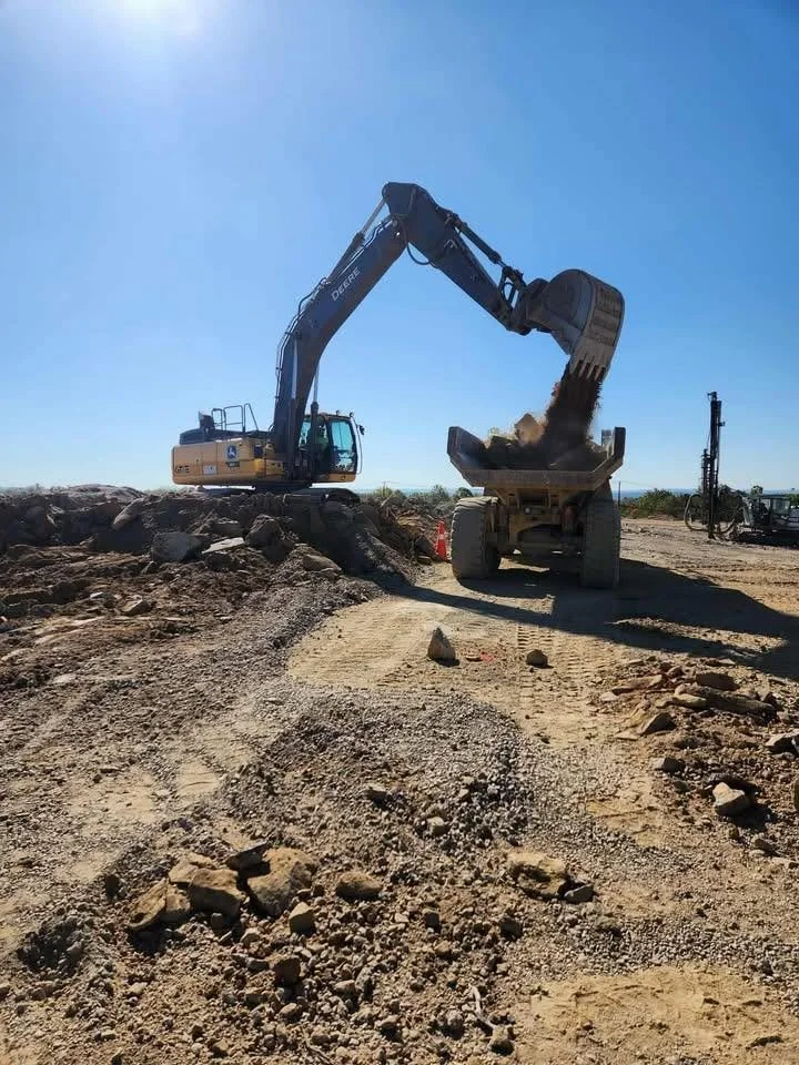 Excavator loading dirt and rocks into a dump truck at a construction site under a clear blue sky.