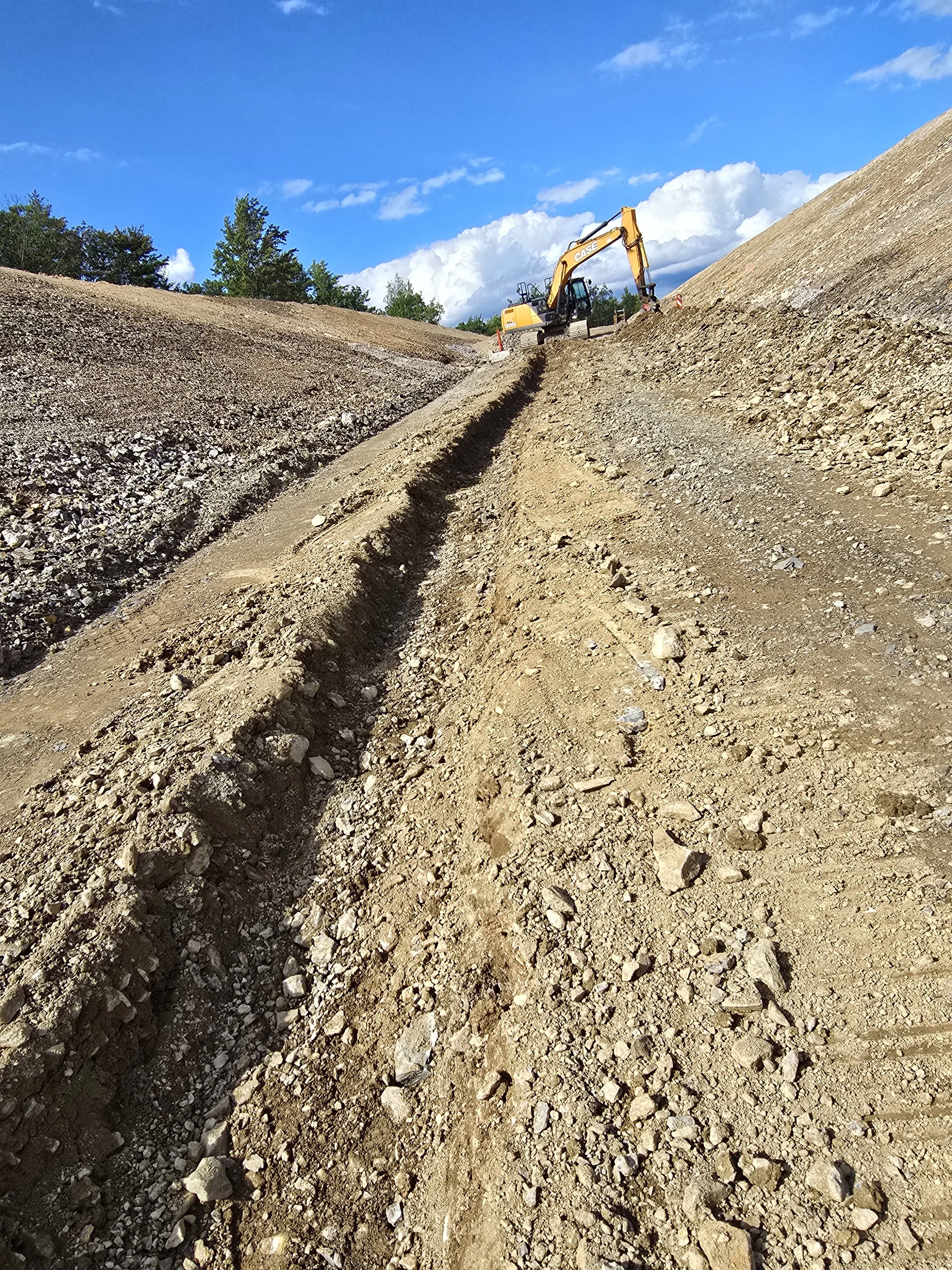 A construction site with a yellow excavator working on a dirt embankment, with trees and a partly cloudy blue sky in the background.