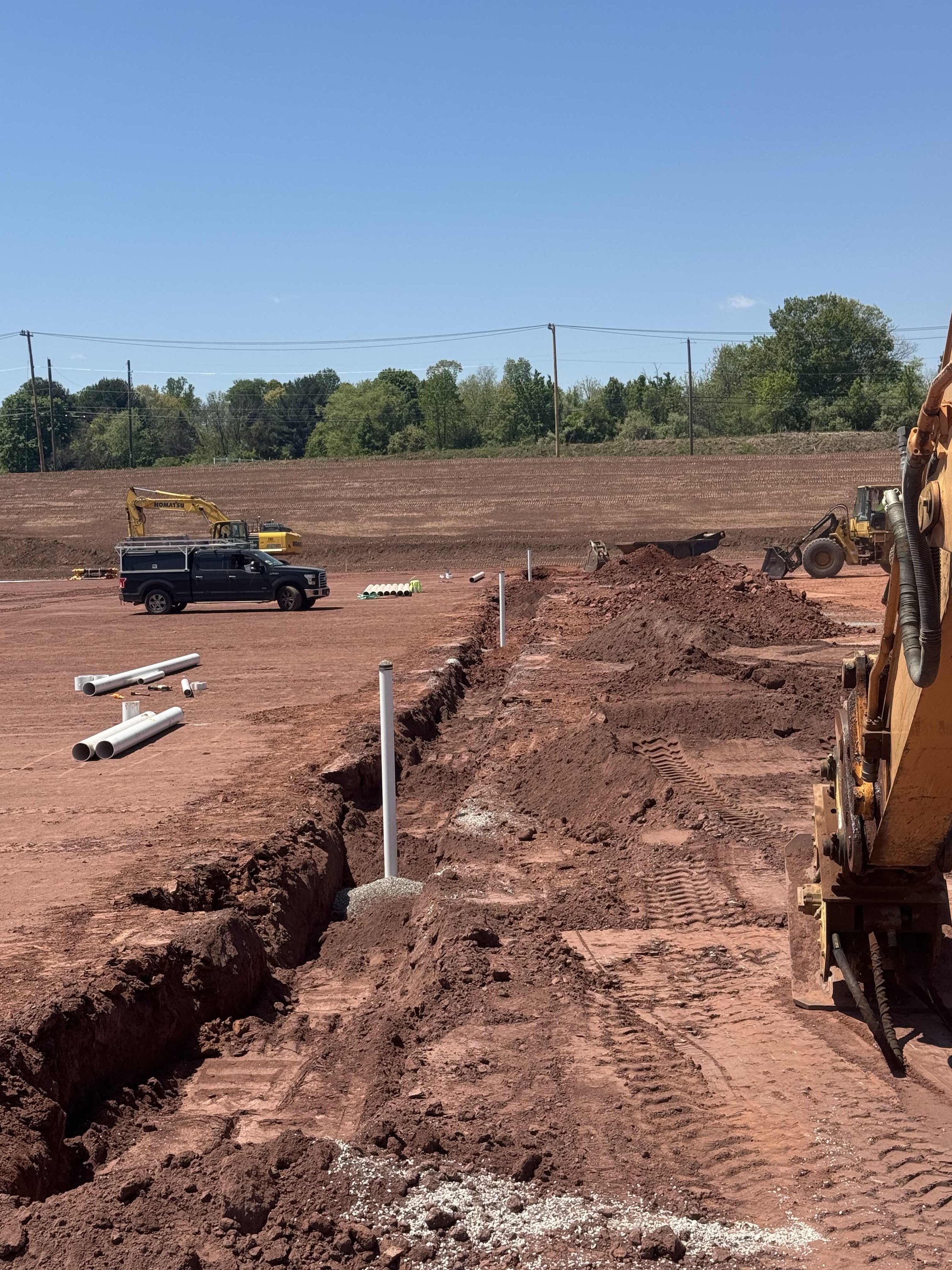 Construction site with dirt trench, some pipes, construction vehicles, and equipment under a clear blue sky.