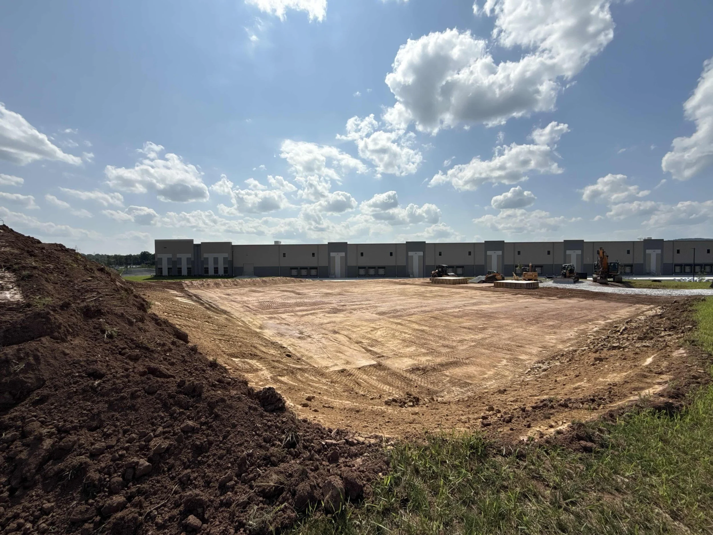 Construction site with a large, gray, commercial building partially built in the background under a blue sky with scattered clouds. Foreground shows disturbed earth and construction equipment.