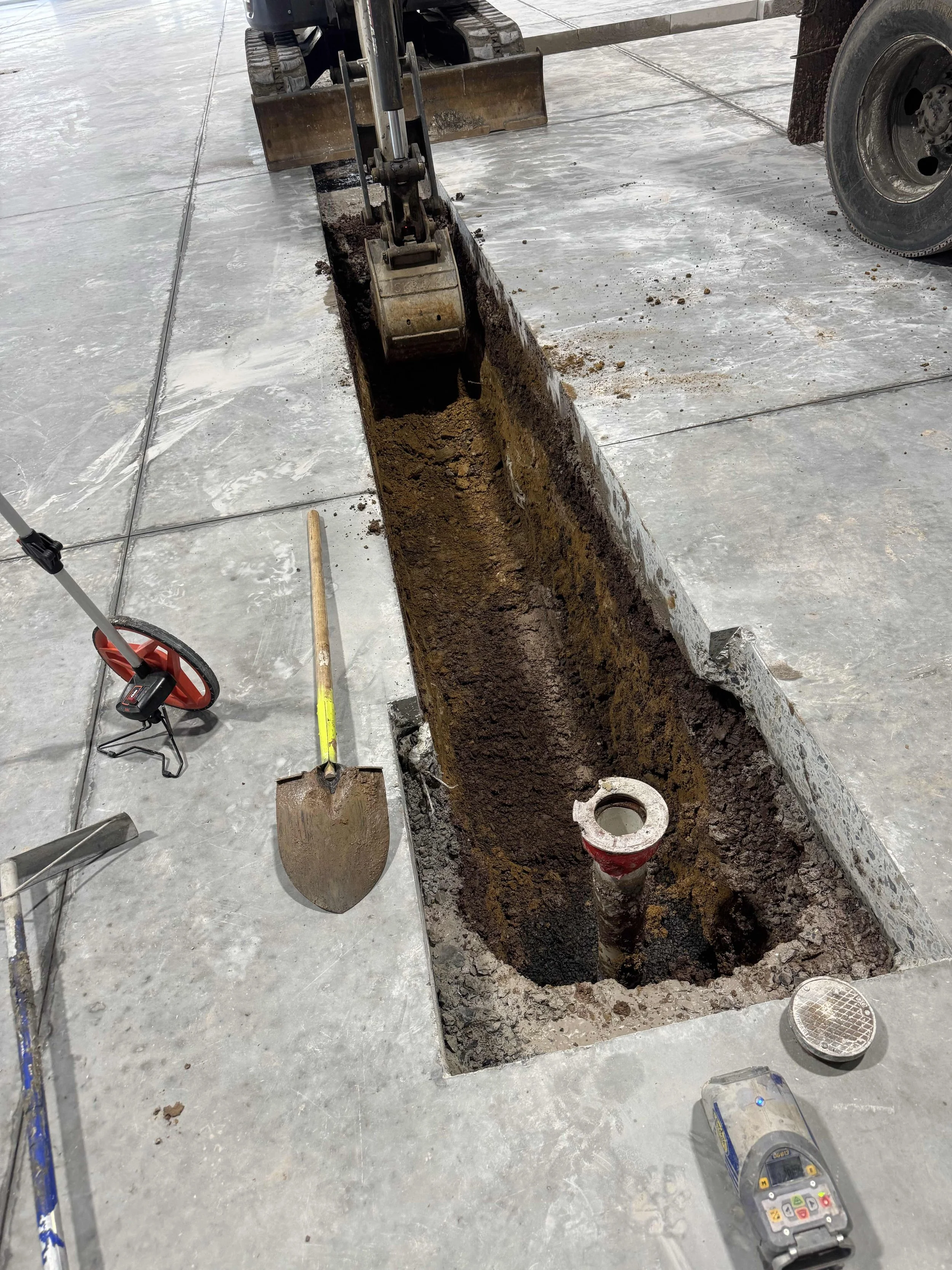A construction site with a rectangular trench in the concrete floor, a shovel resting on the ground nearby, and construction tools including a level, a cordless drill, and a power saw placed around the trench. A small excavator and a tire are visible