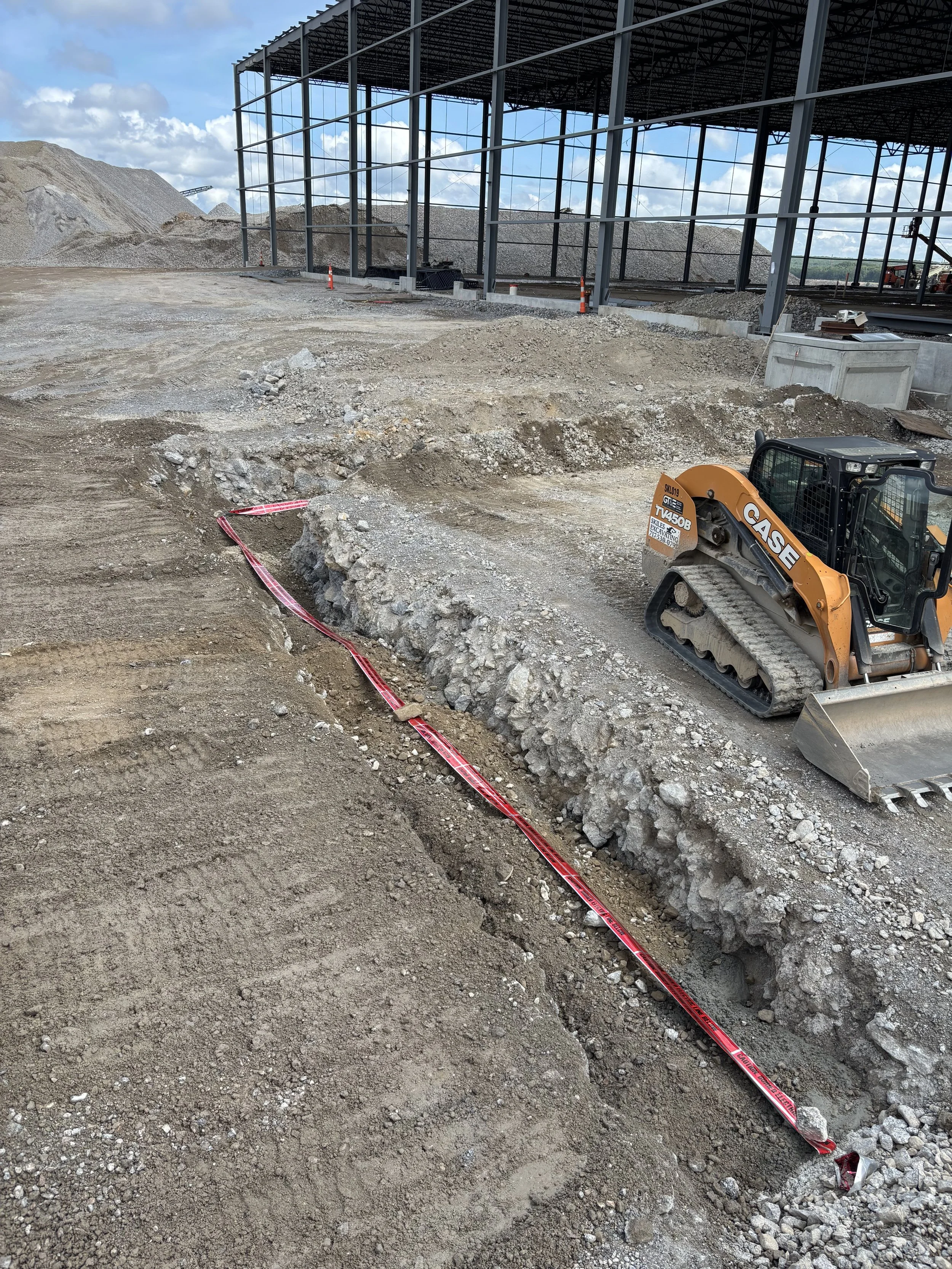 Construction site with dirt and gravel, a small excavator, a large steel structure, and piles of dirt against a partly cloudy sky.