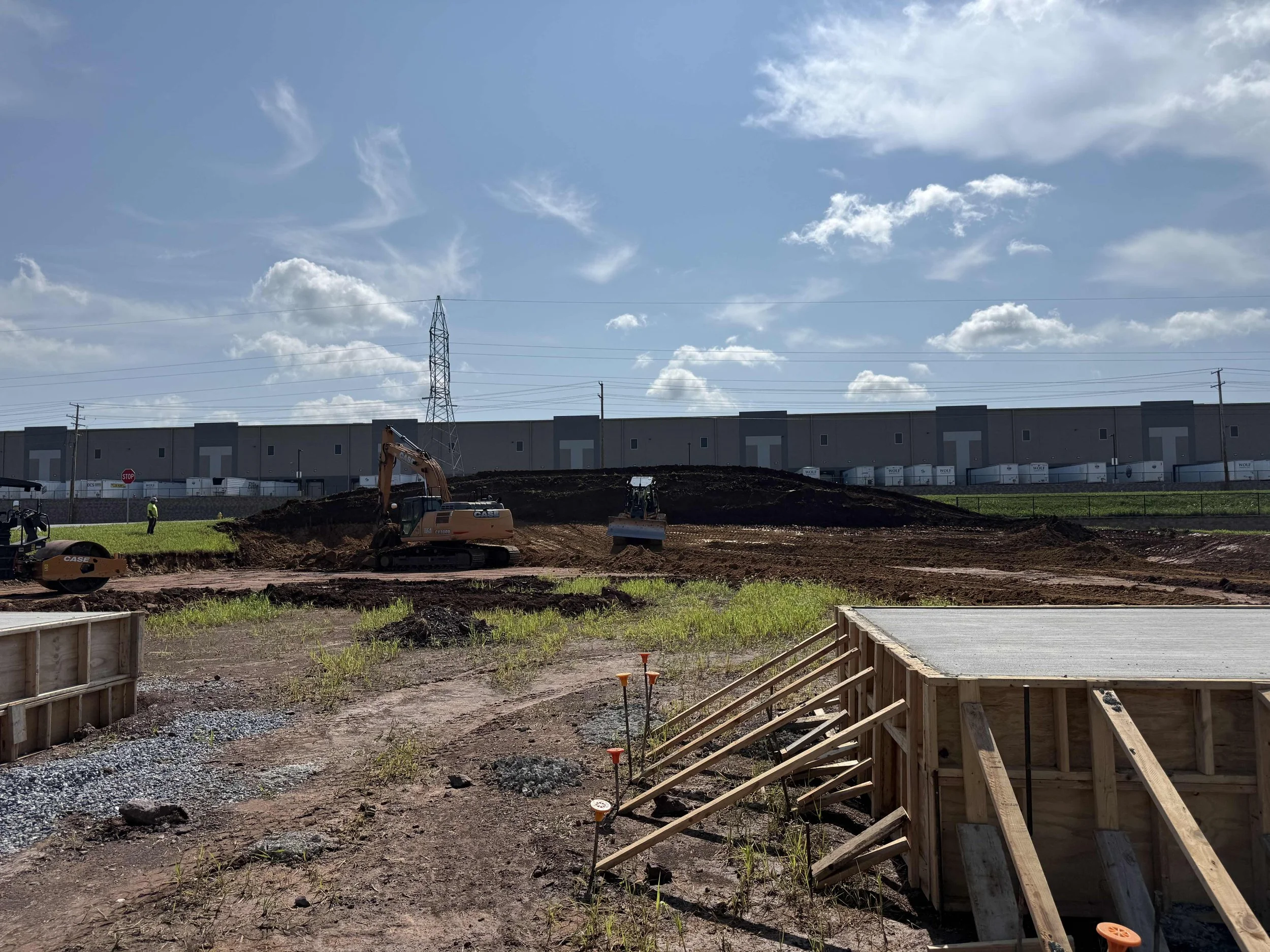 Construction site with machinery, workers, and foundation work underway. A large warehouse building is visible in the background under a partly cloudy sky.