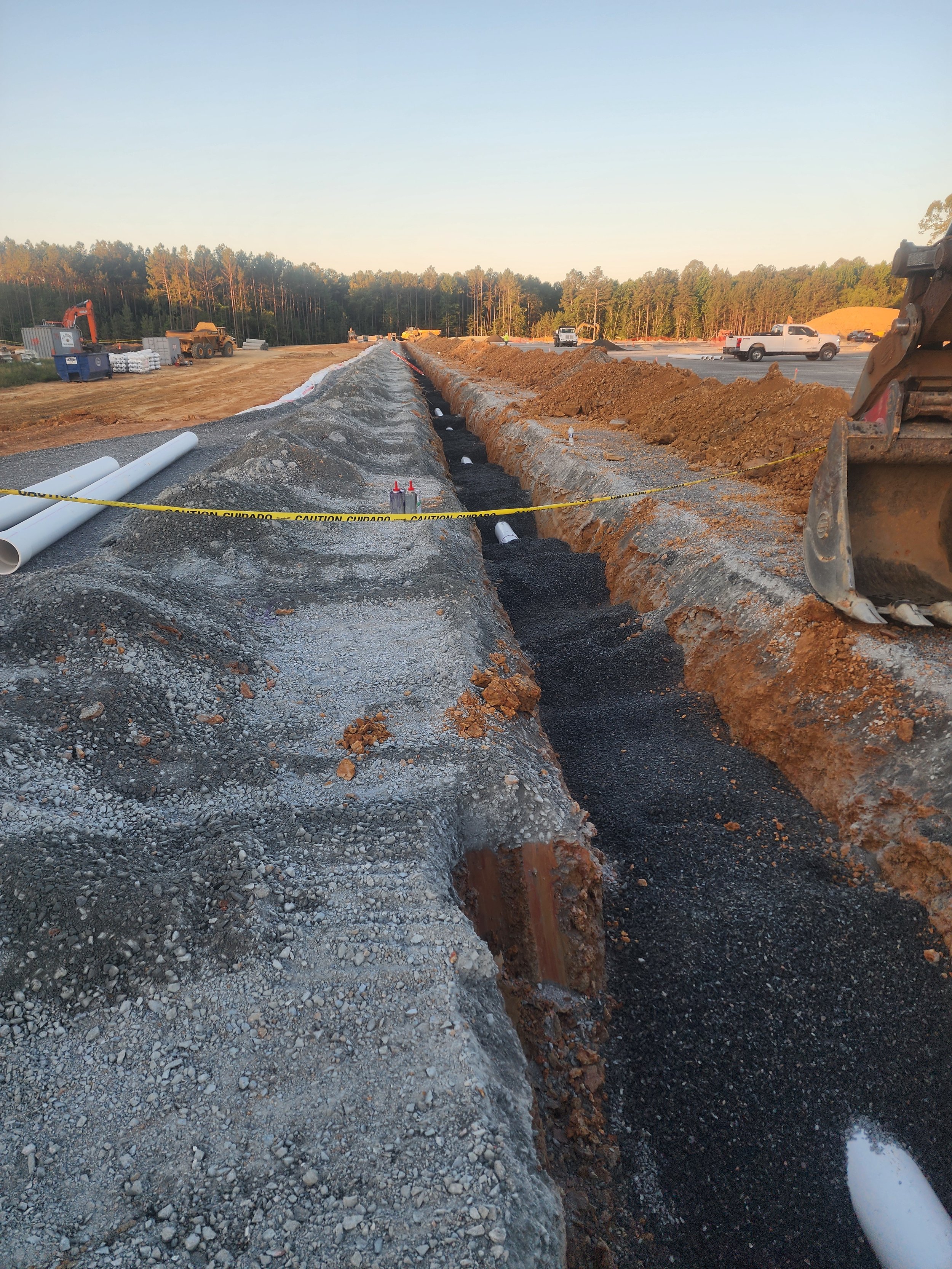 Construction site with a trench for underground pipes, dirt piles, construction equipment, and vehicles, with caution tape and pipes on the ground.