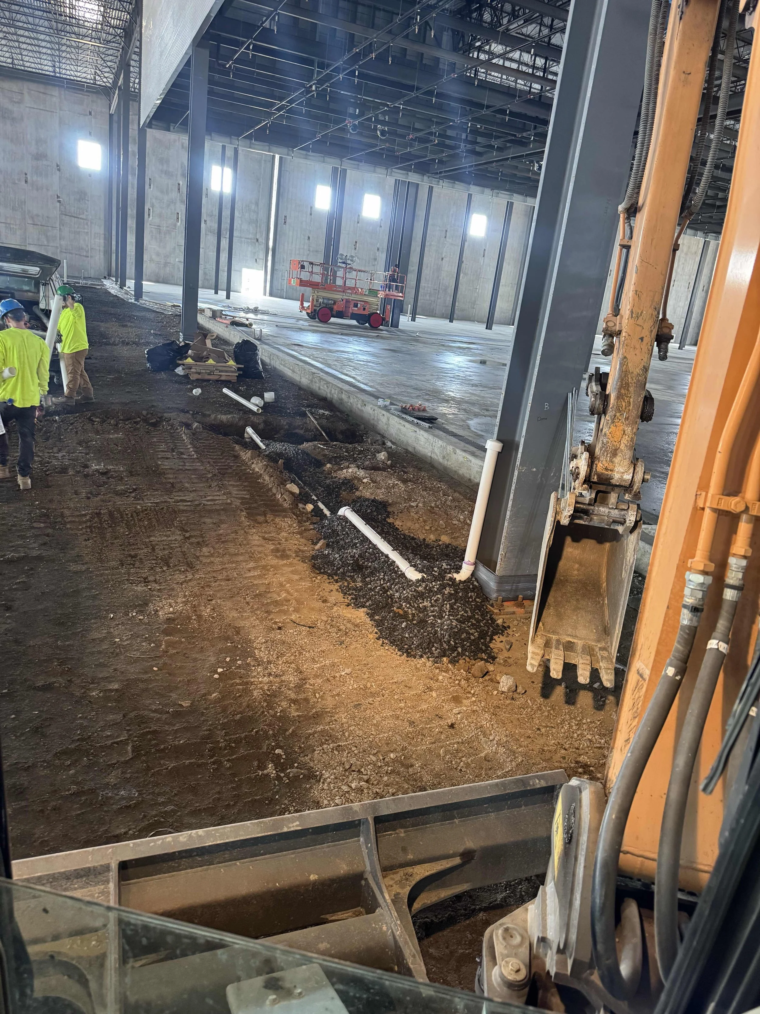 Construction workers in high-visibility clothing working inside a large industrial building under construction. The interior features steel beams, a concrete floor, and construction equipment, including an excavator and a scissor lift.