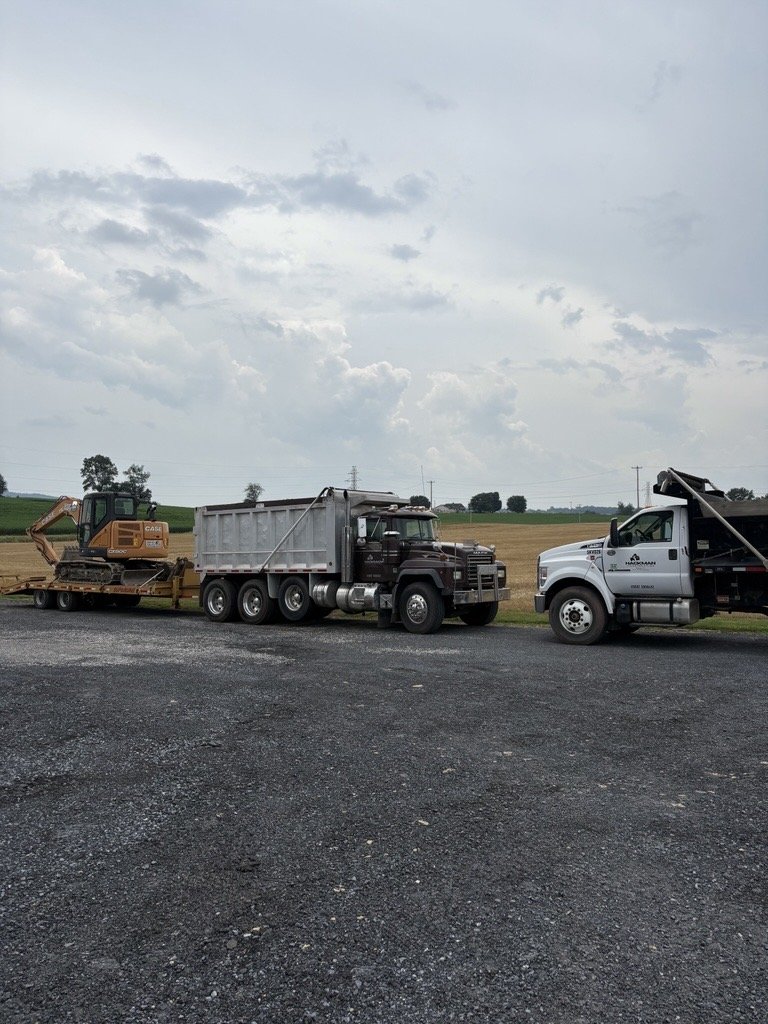 A construction site with a dump truck, a pickup truck, and a flatbed trailer carrying a small excavator on a rural road, under an overcast sky.
