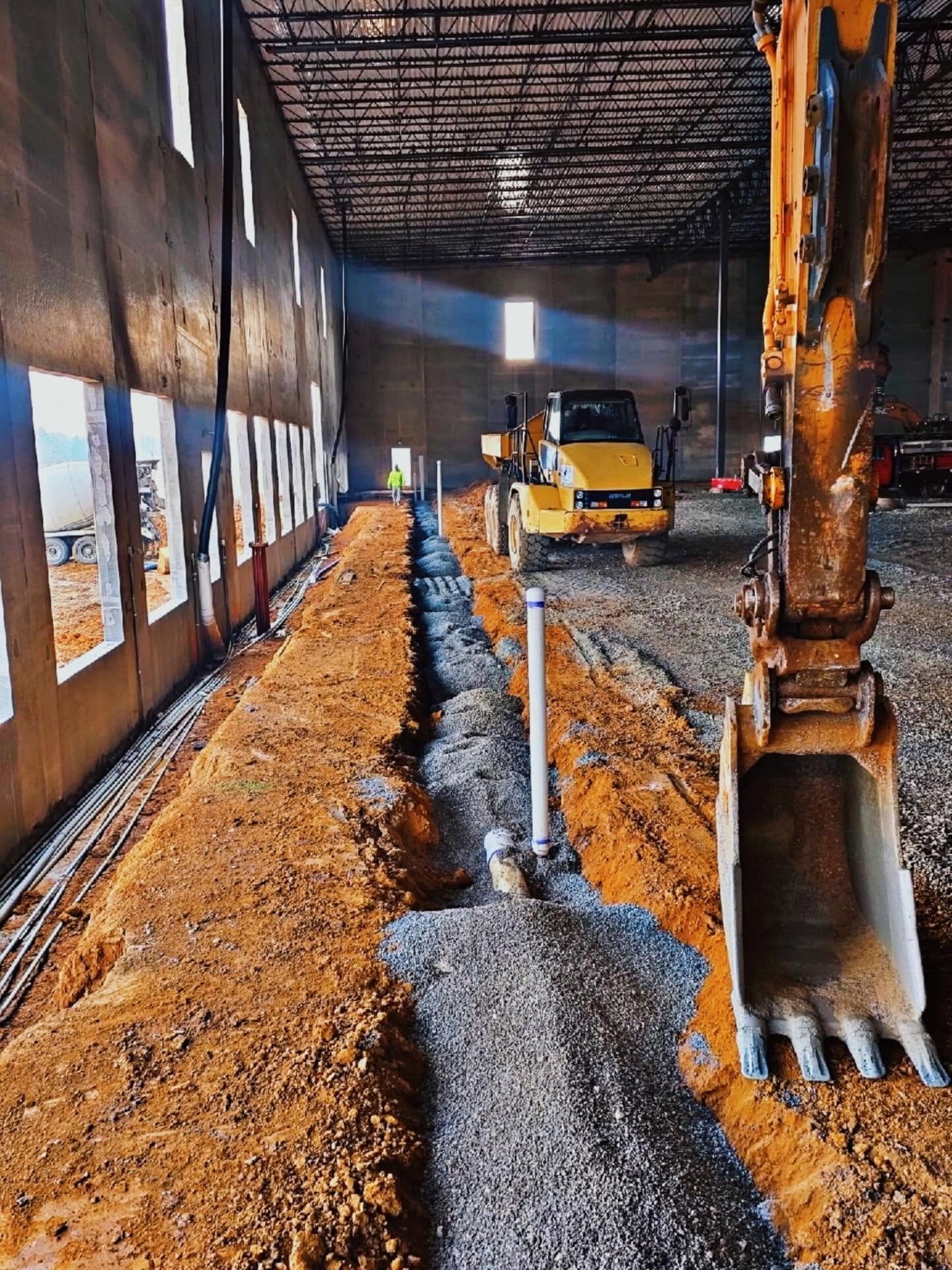 Construction site inside a large building with a yellow bulldozer, gravel, and an excavator arm visible in the foreground. Workers in the background are wearing high-visibility clothing. The building has a high ceiling with metal framing and windows,