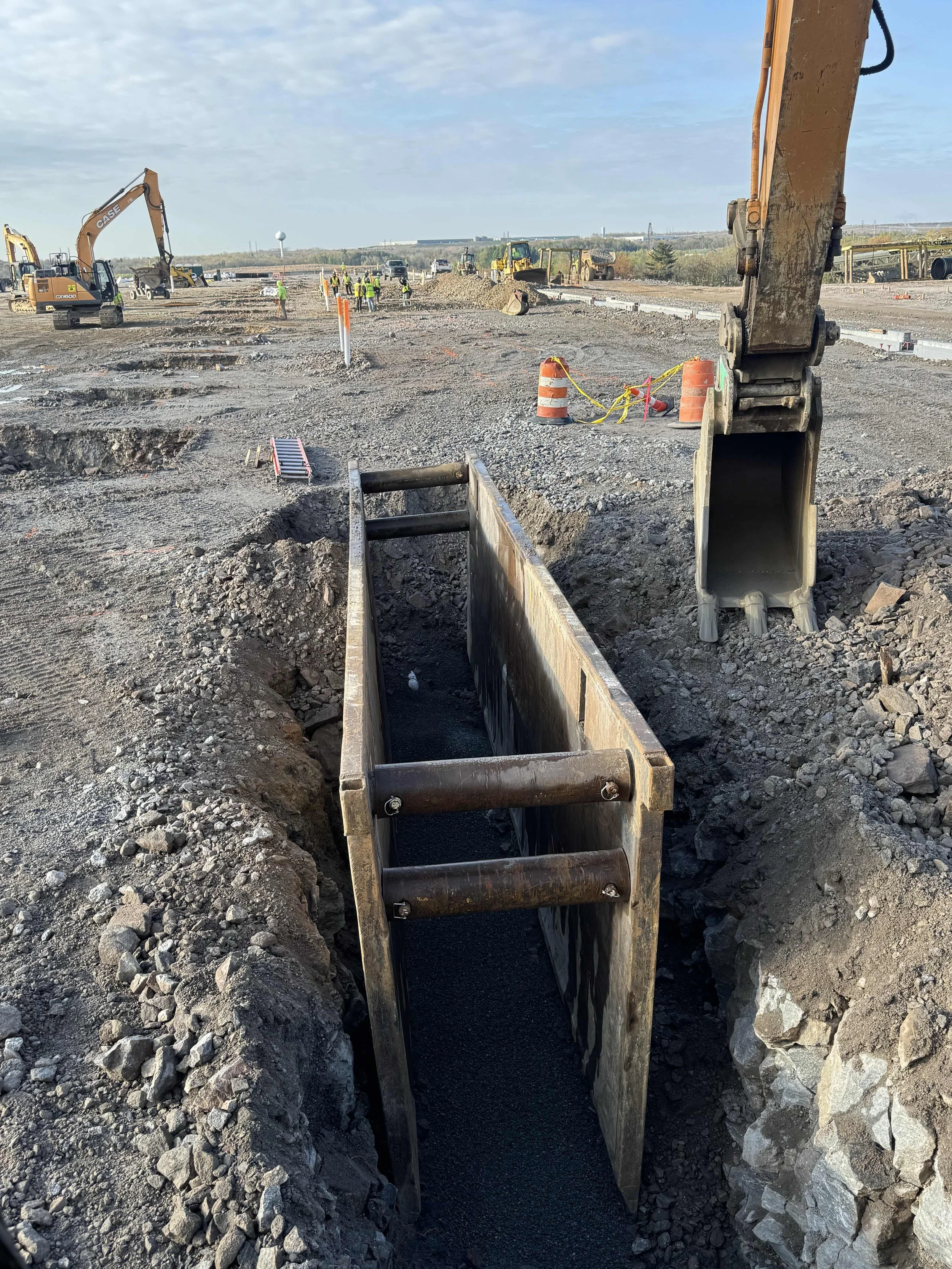 Construction site with heavy machinery and workers, trench being dug, and a clear blue sky in the background.