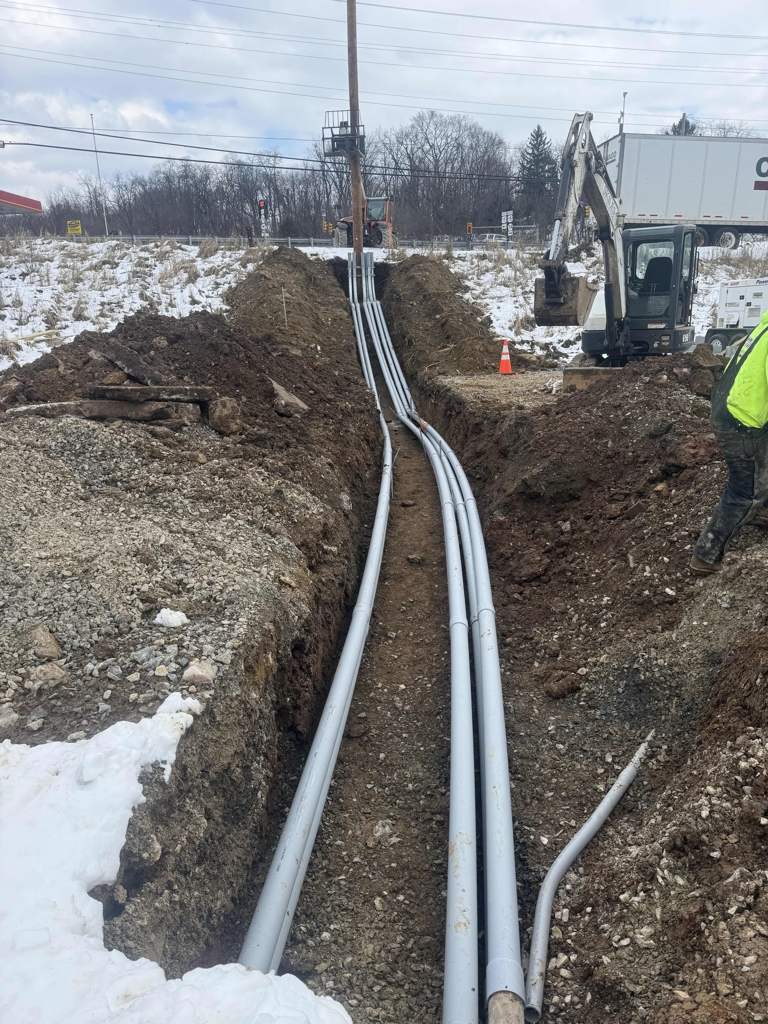 Construction workers installing electrical or communication conduits in a trench dug in snowy ground, with equipment and a small excavator present.