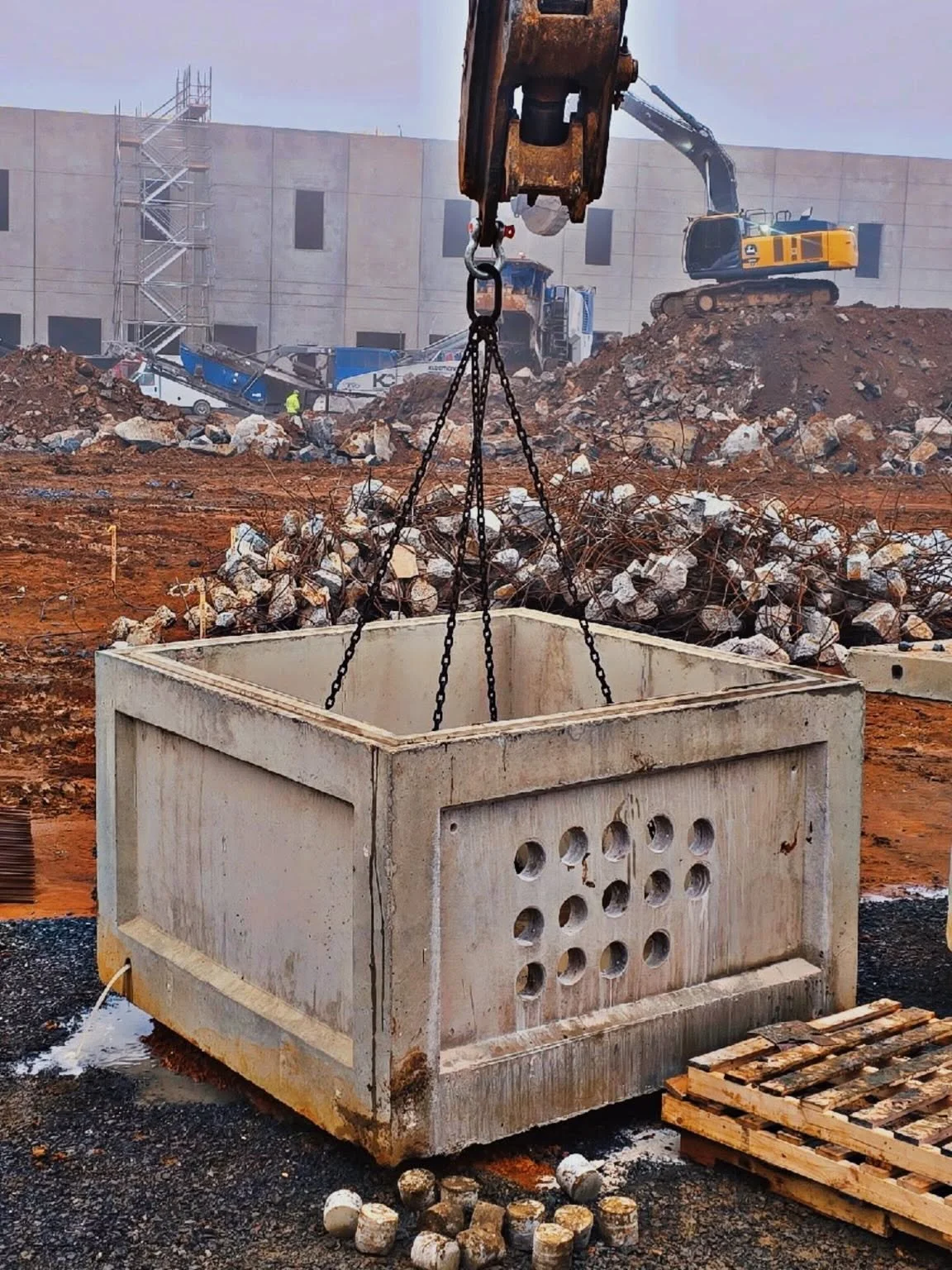 A construction site with excavators, piles of rocks and debris, and a large concrete container being lifted by a crane.