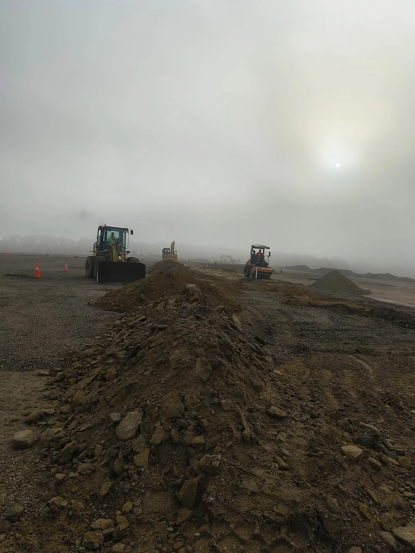 Construction site with dirt piles, excavators, and orange traffic cones, under a cloudy sky with the sun visible through the fog.