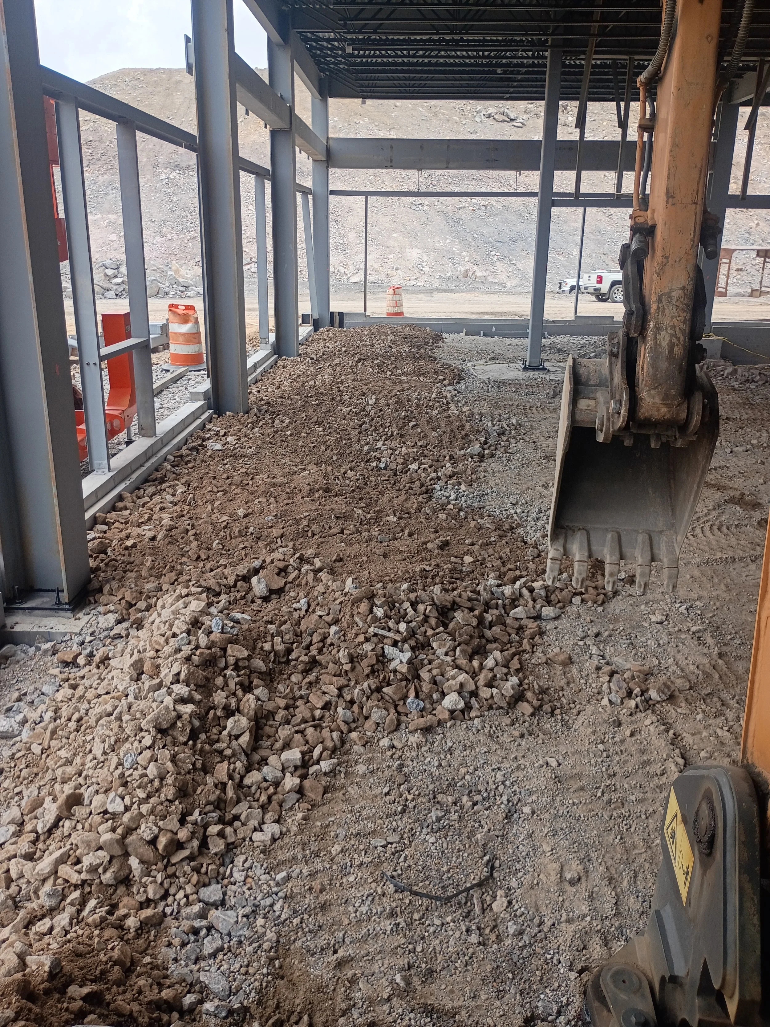 Construction site with an excavator, dirt, rocks, orange traffic cones, and a partially built structure with metal framing.