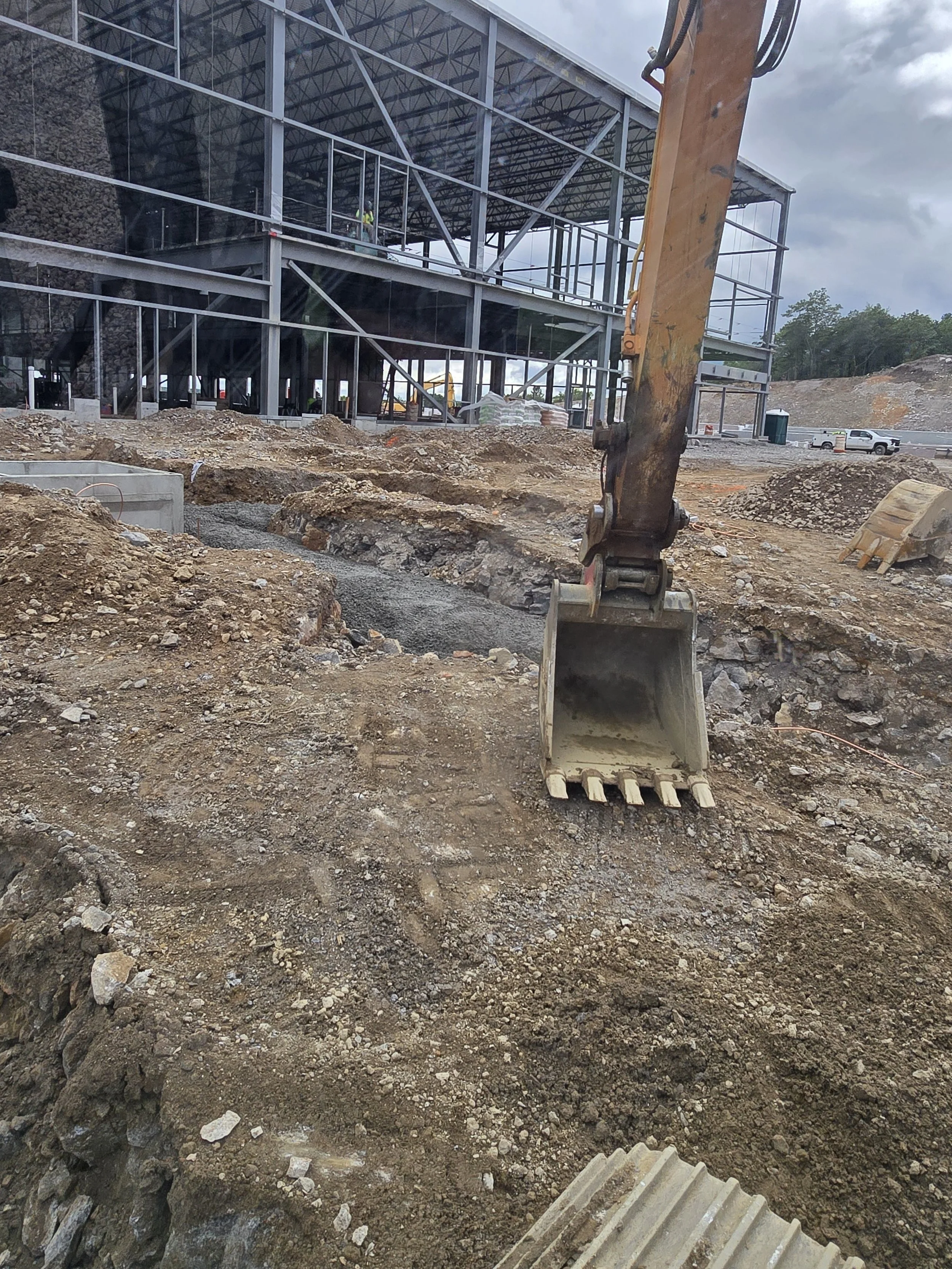 Construction site with a large excavator bucket in the foreground, ongoing earthwork and a steel framework building under construction, cloudy sky in the background.