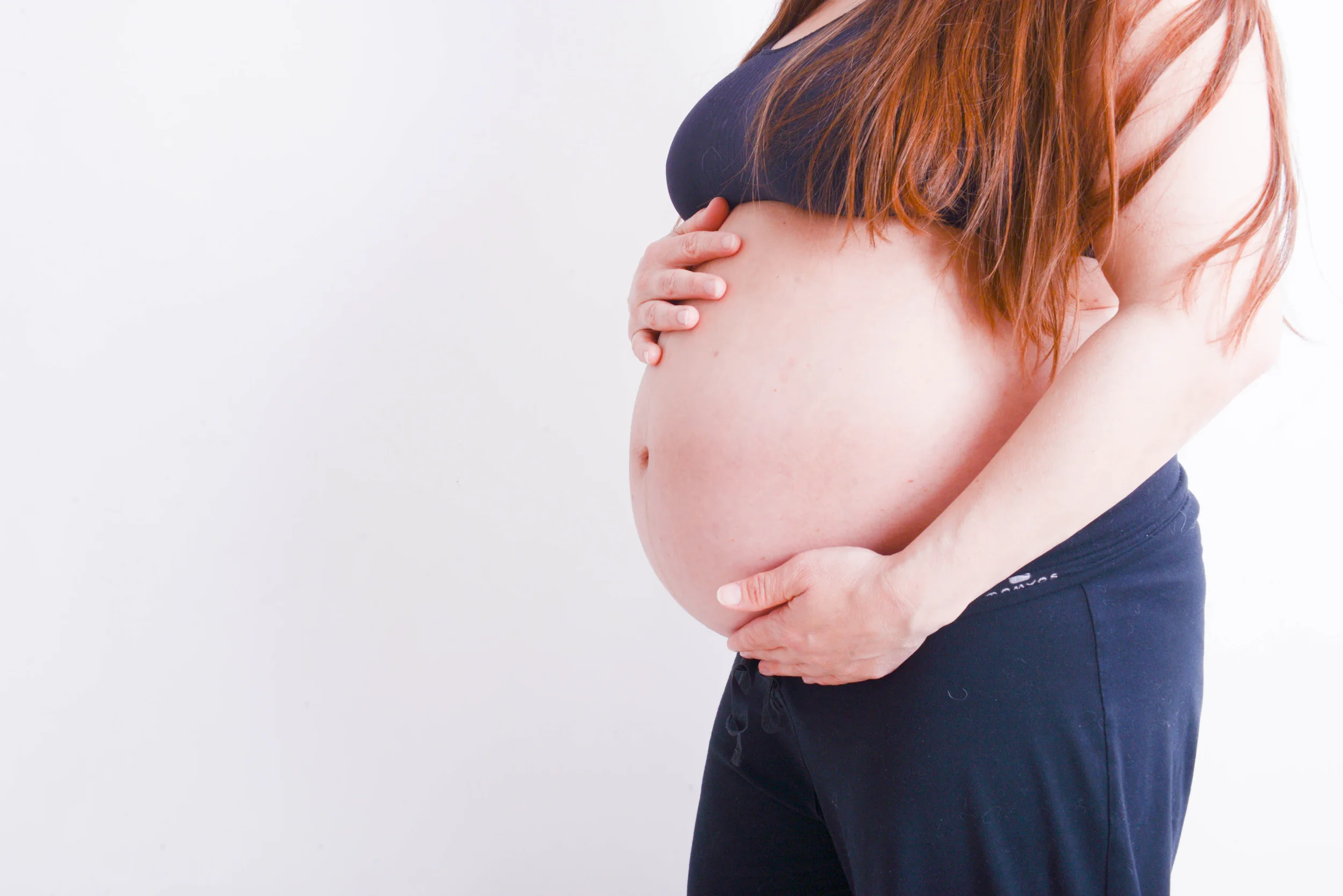 A pregnant woman with red hair in a dark sports bra and dark pants holding her belly with both hands against a white background.