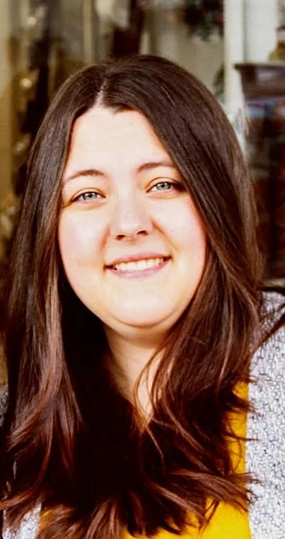 A young woman with long brown hair, smiling, with a blurred outdoor background.