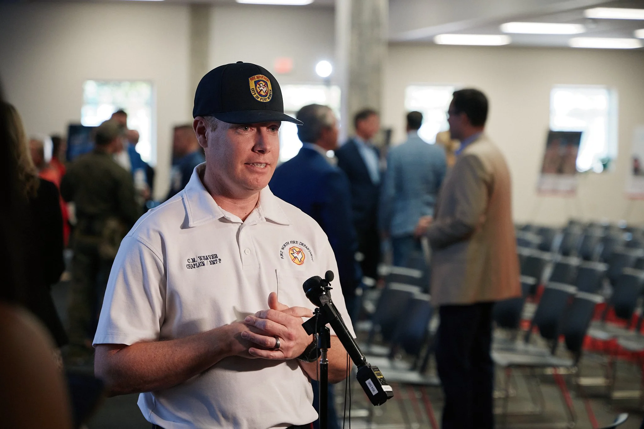 A man wearing a white shirt with a fire department logo and a baseball cap, standing in front of a microphone, speaking to an audience at an indoor event.