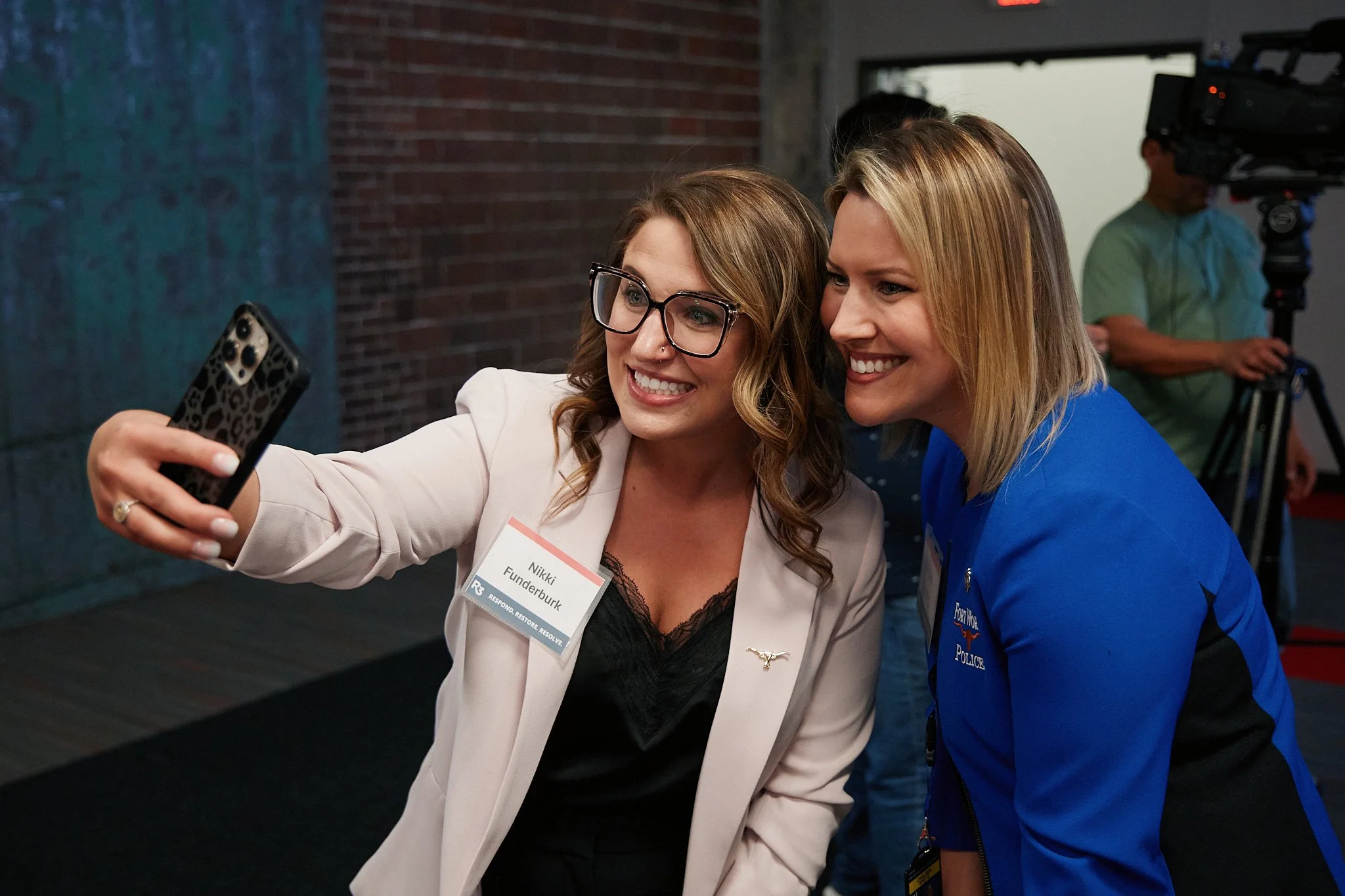Two women are taking a selfie together while smiling. One woman is wearing glasses and a white blazer, and the other is wearing a blue uniform with a police badge. They appear to be at an indoor event with a cameraman in the background.