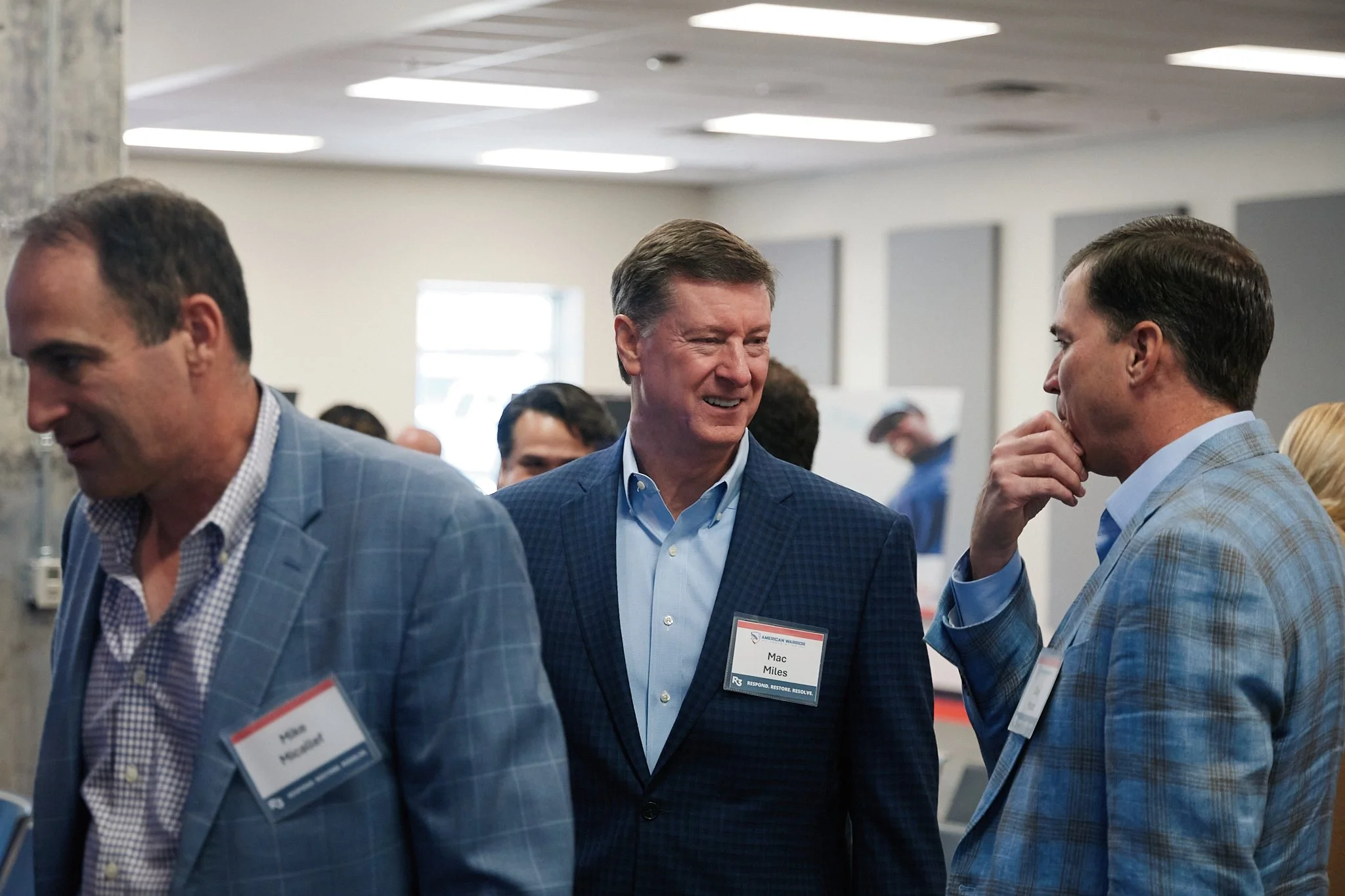 Three men in business attire engaged in conversation at a professional event or conference, with name tags visible.