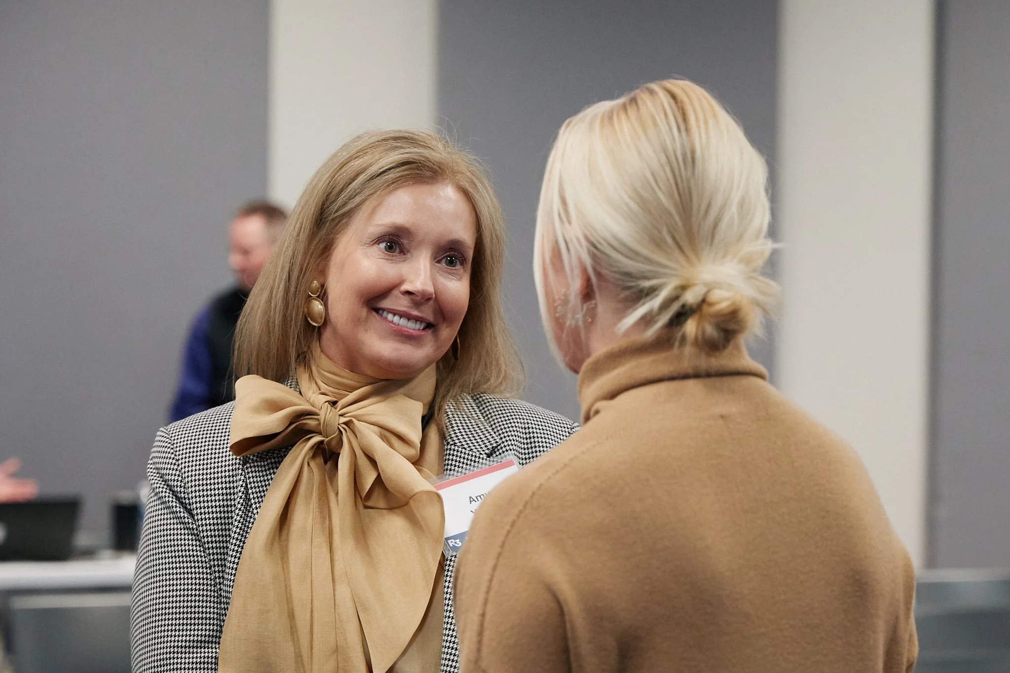 Two women engaged in conversation at a professional event, one woman with blonde hair wearing a checkered blazer and beige scarf, the other with light blonde hair in a bun wearing a tan coat.