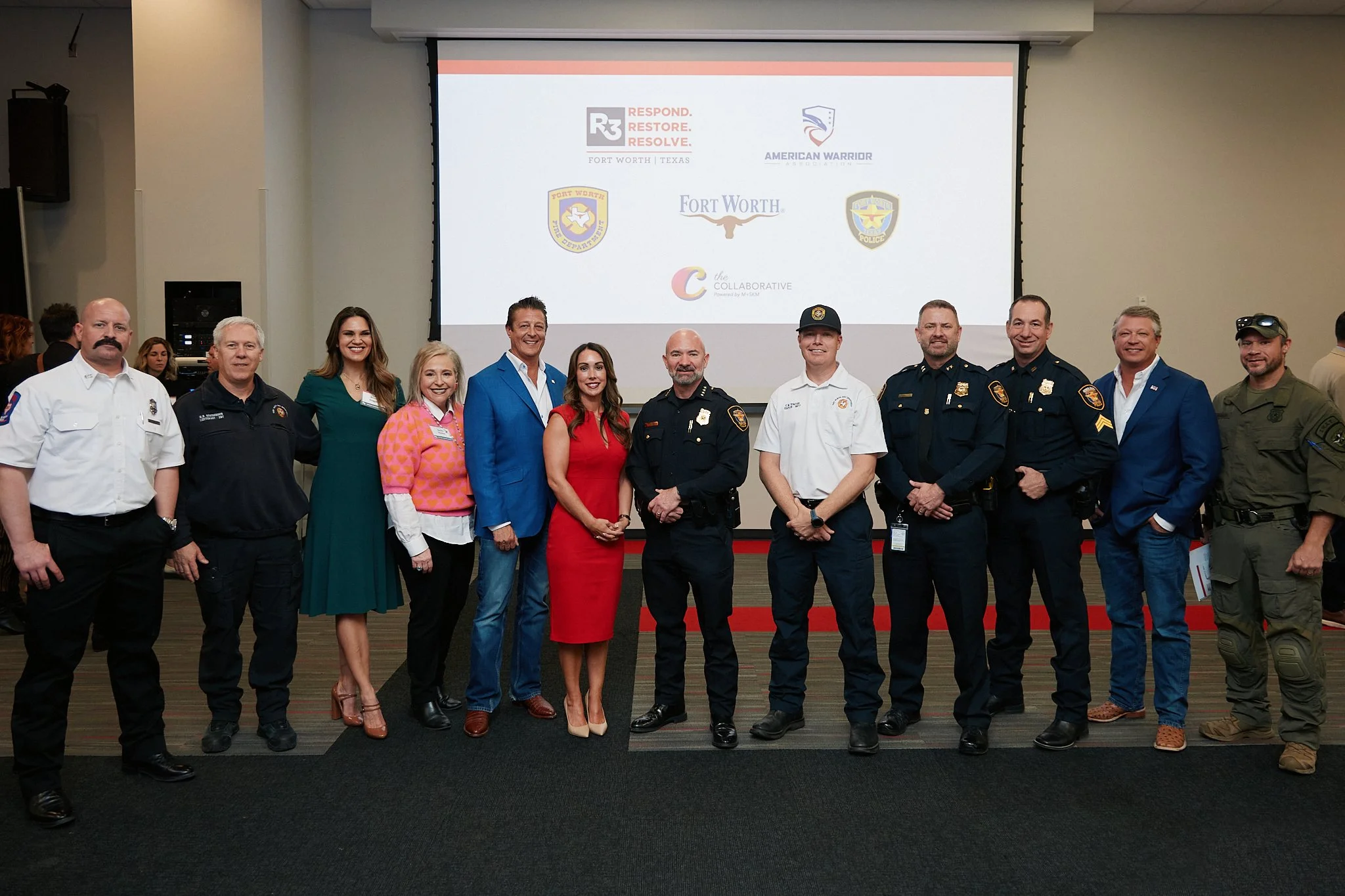 A group of people, including police officers, emergency responders, and civilians, standing side by side in front of a presentation screen with various logos, in a conference or event setting.