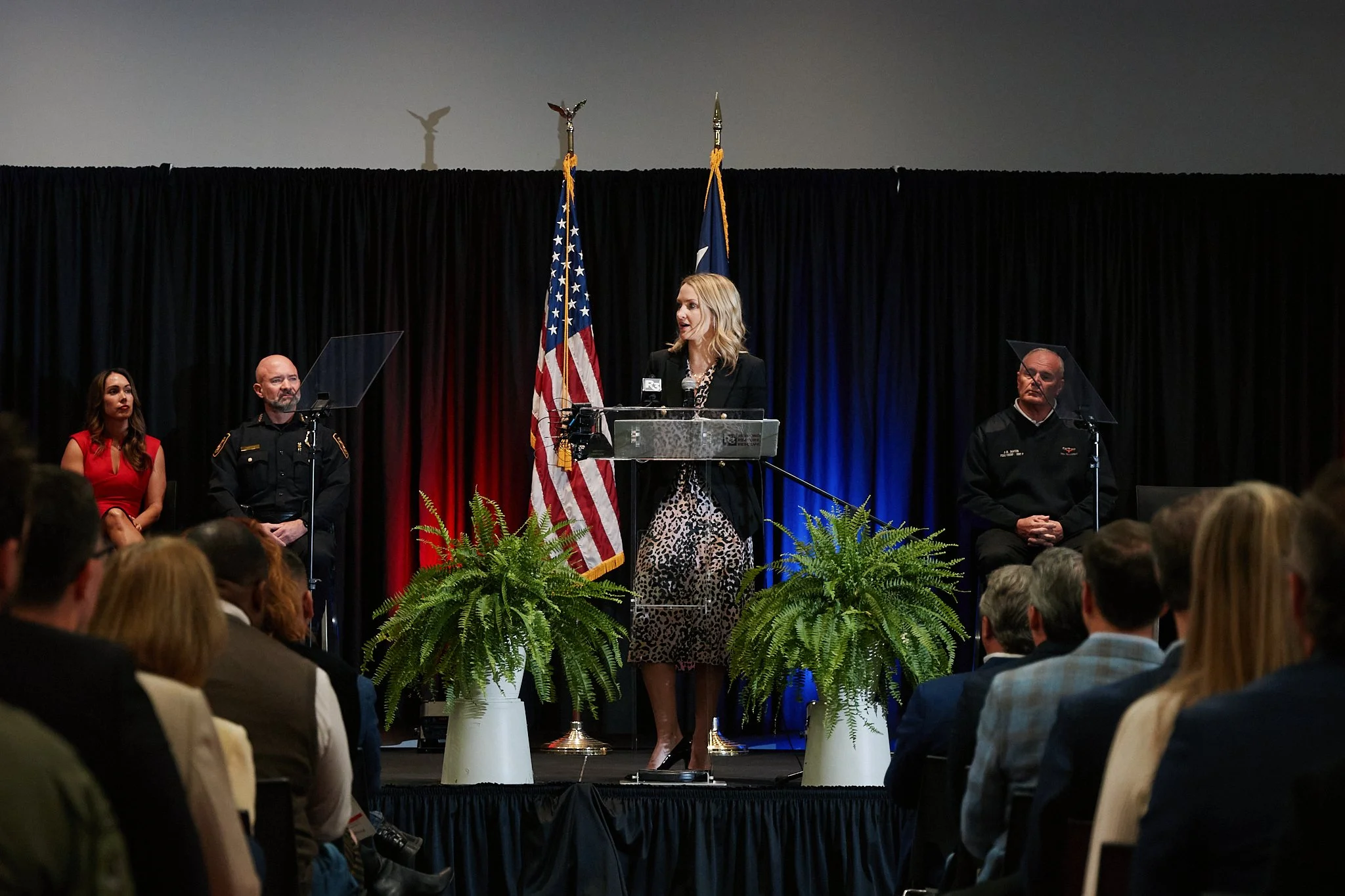 A woman giving a speech at a podium on stage with American flag and a blue backdrop, flanked by three individuals seated behind acrylic shields, and a gathered audience in front.