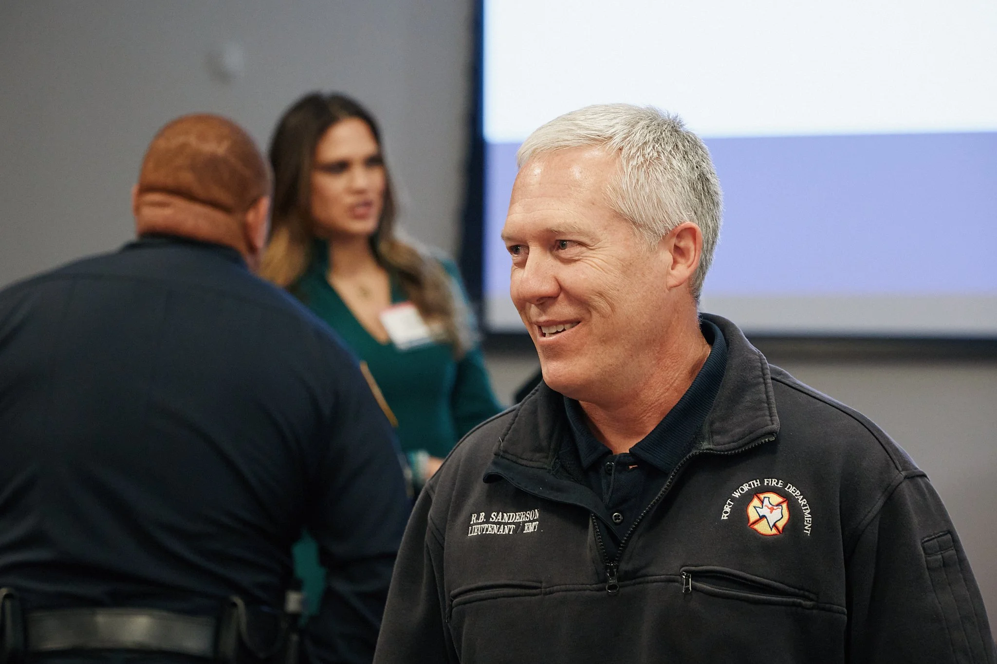 A man with gray hair smiling in a black uniform with a patch that reads "Port Worth Fire Department" and a name tag that says "R.B. Sanderson" talking to a person with a bald head in a dark uniform, with a woman with long hair in the background