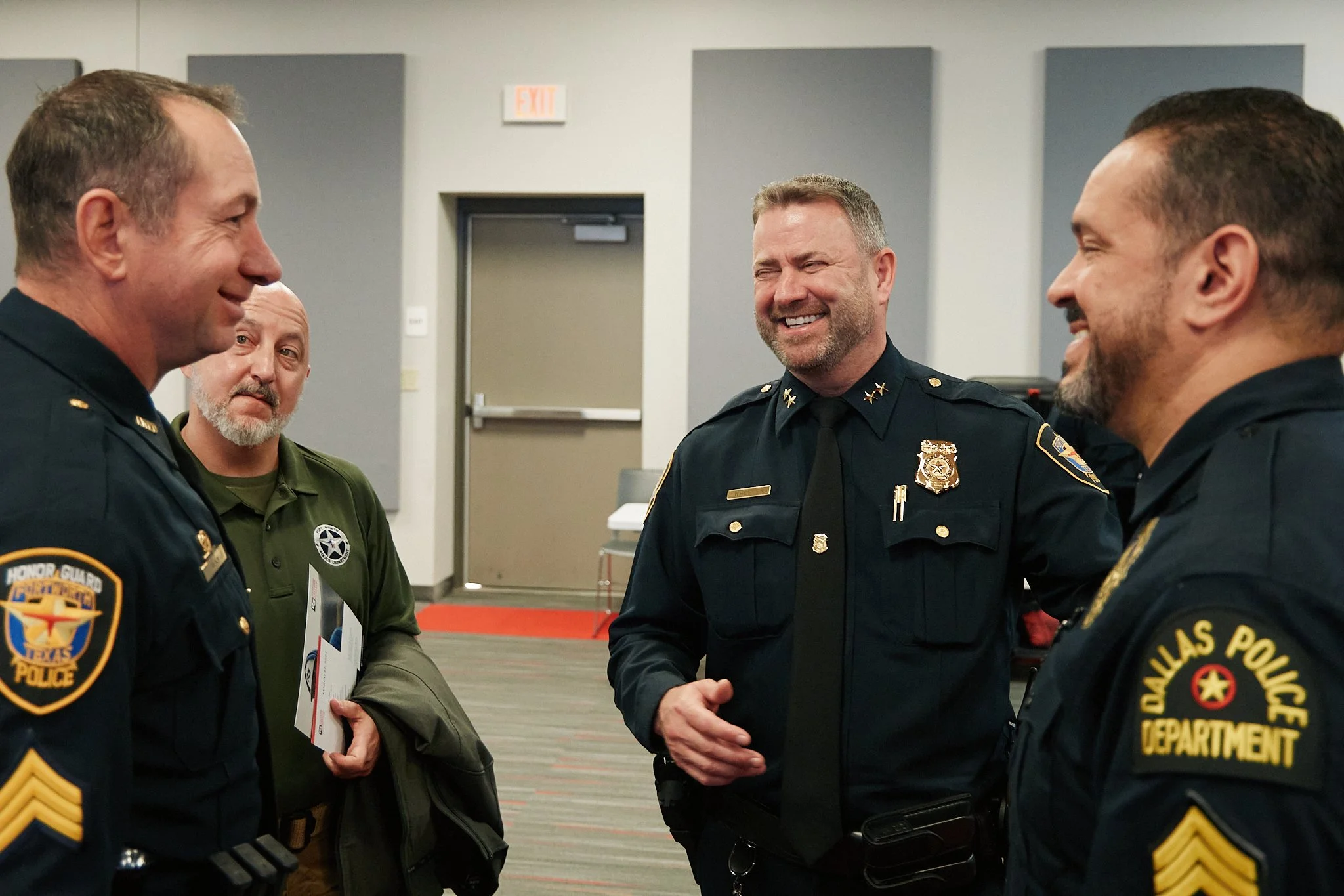 Group of four police officers talking and laughing indoors with gray walls and a door in the background.
