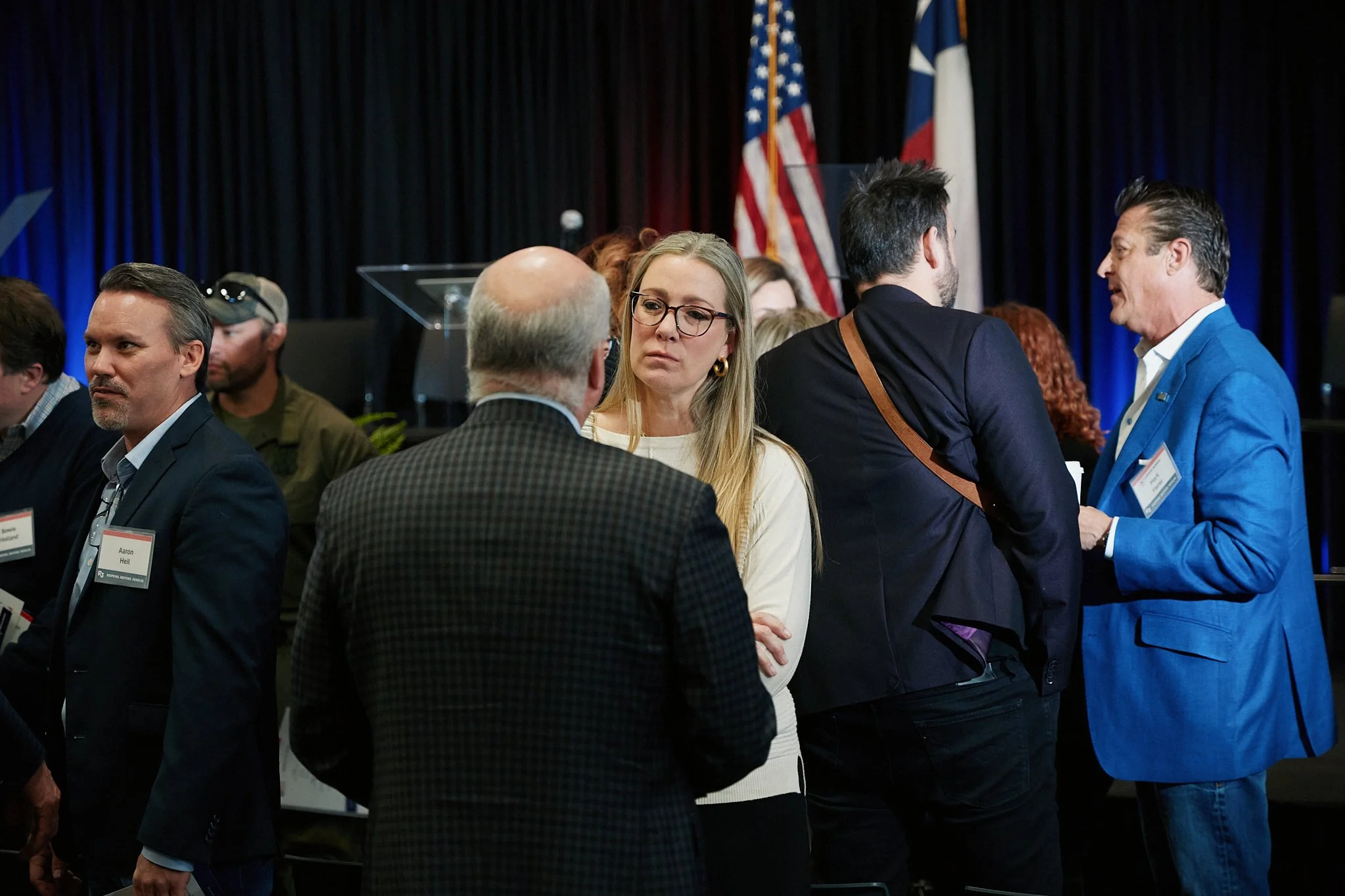 People engaged in conversation at a political or professional event with American and Texas flags in the background.