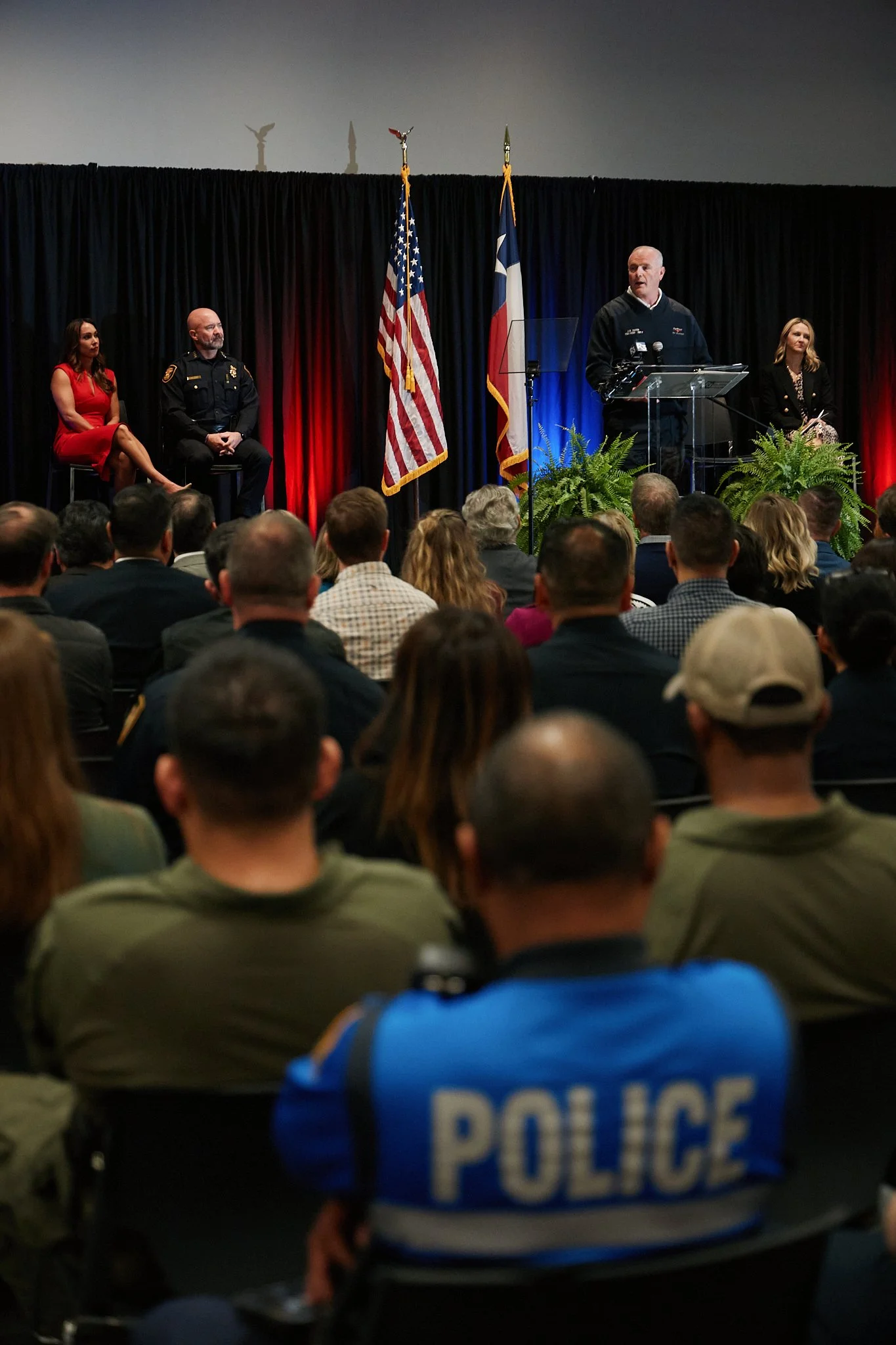 A man giving a speech at a podium during a formal event with an audience, two women, and a police officer on stage, with American and Texas flags behind them.