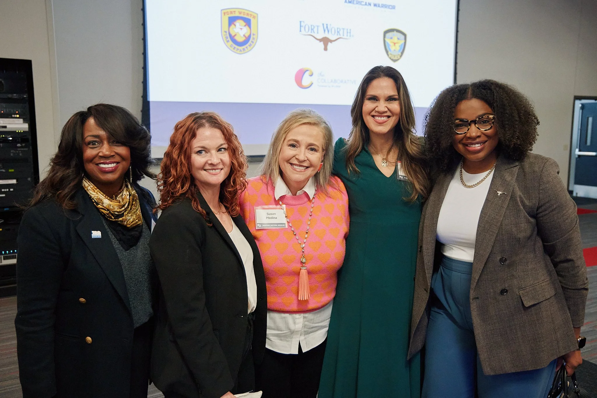 A group of five women standing together smiling at a conference or event, with a screen showing logos in the background.