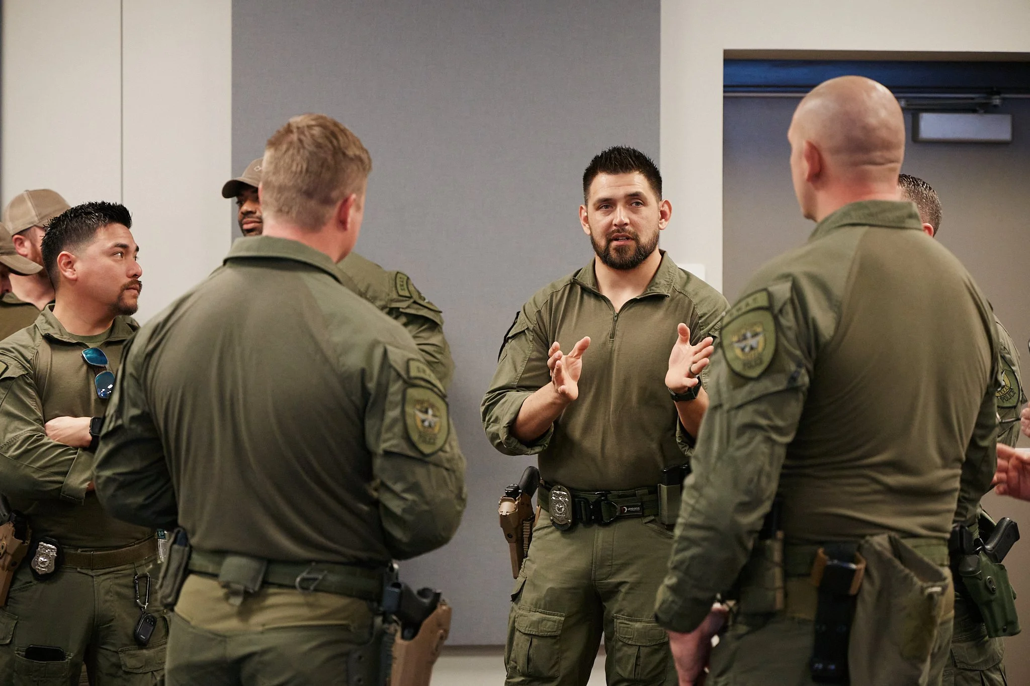 A group of law enforcement officers in uniform, gathered in a room and listening to a man who appears to be giving instructions or a briefing.