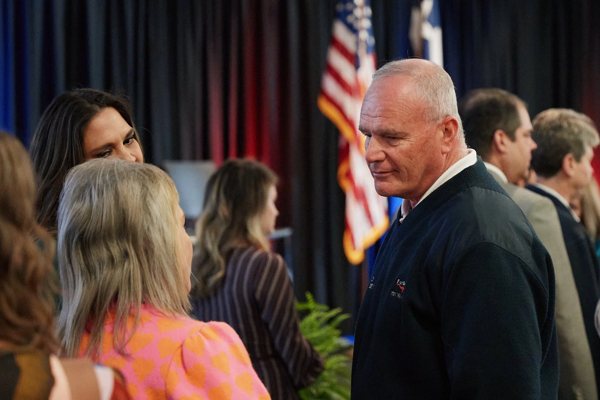A man with short white hair in conversation with a woman with blonde hair, among other people, on a stage with American flags and dark curtains in the background.