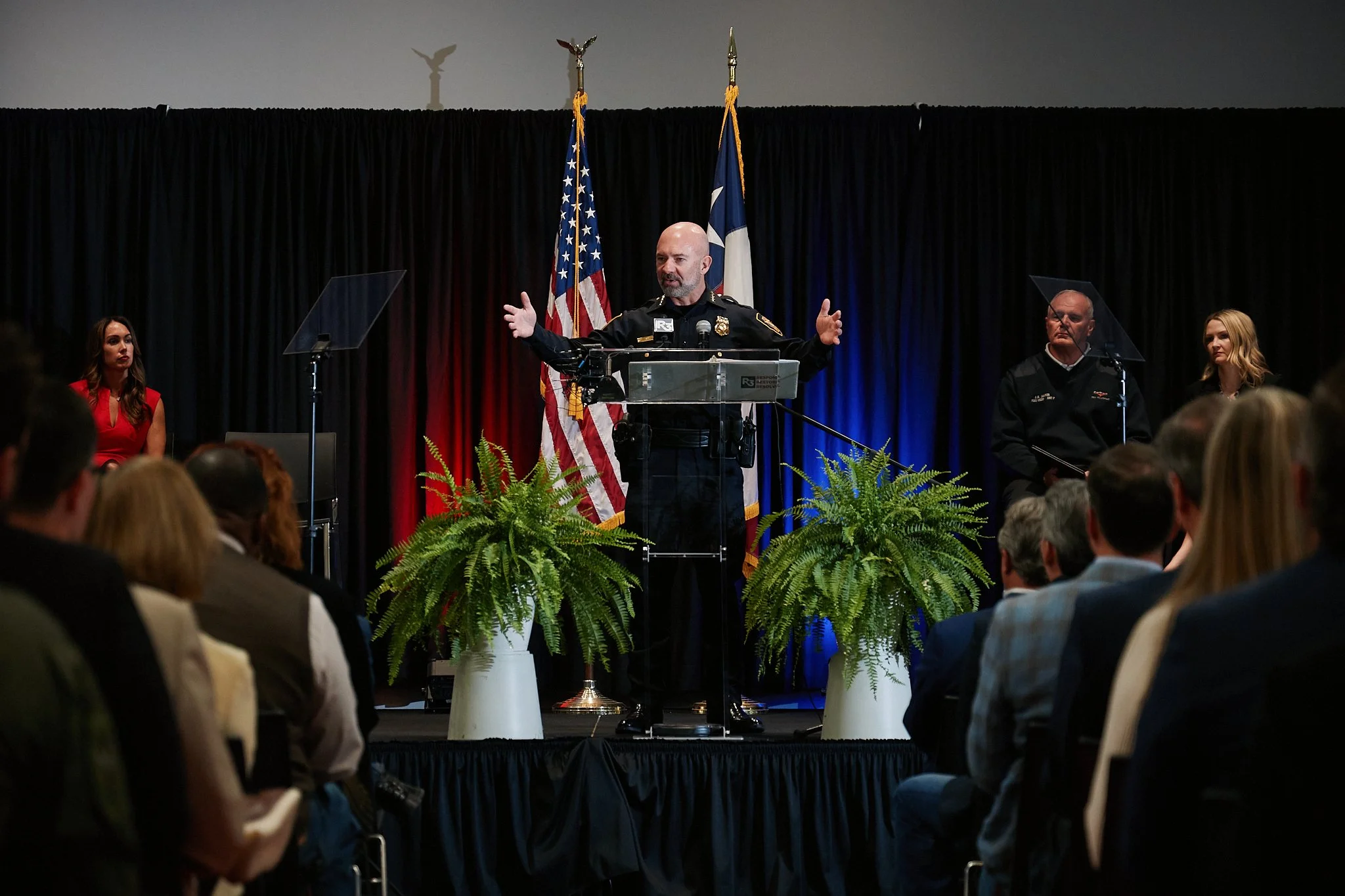A police officer giving a speech at a podium during an indoor event with an audience. There are two American flags and a blue and red curtain backdrop. The audience is seated and listening attentively.
