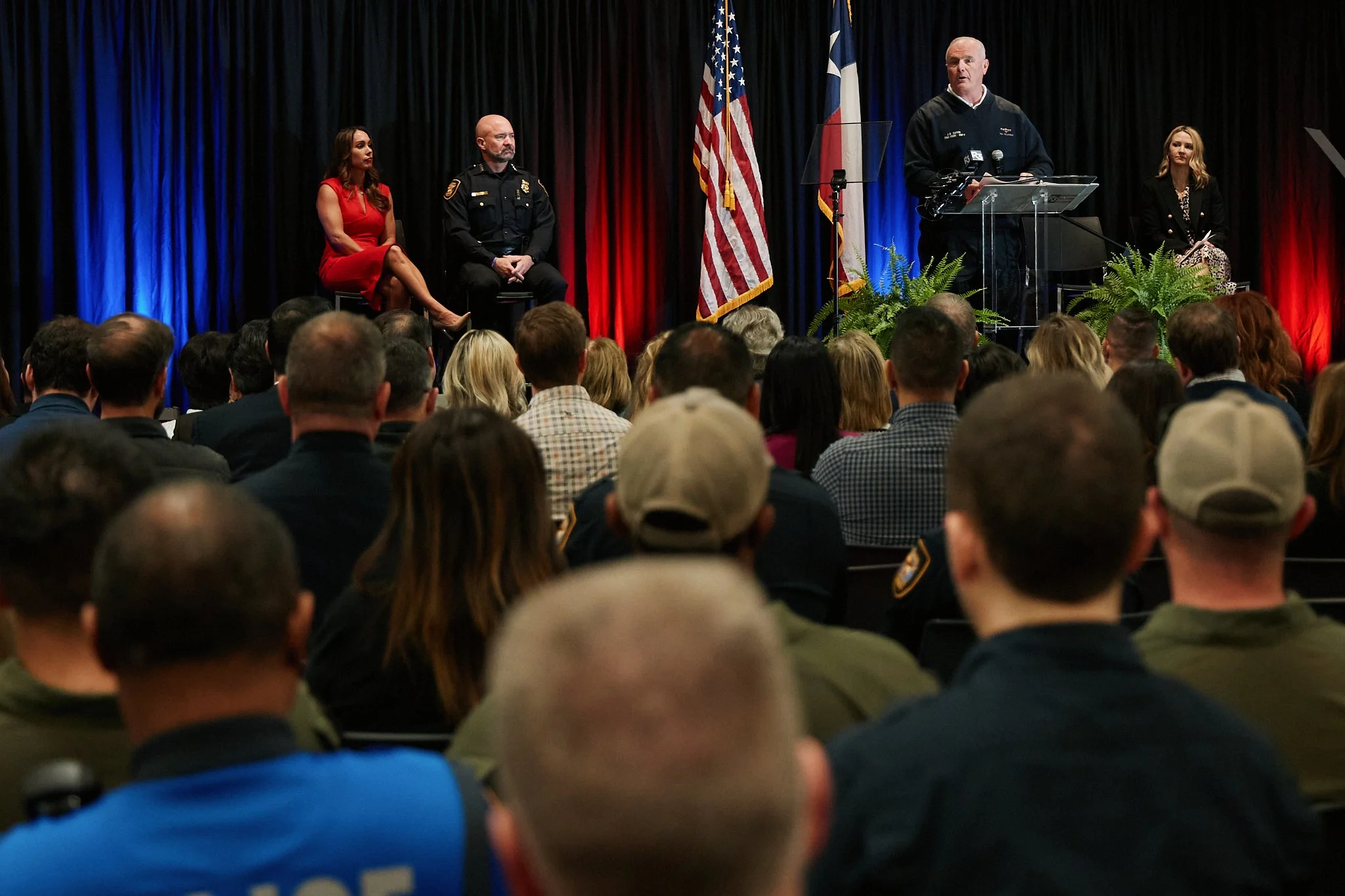 A man at a podium speaking during a public event with an audience of police officers and civilians, U.S. flags, and a black, red, and blue curtain backdrop.