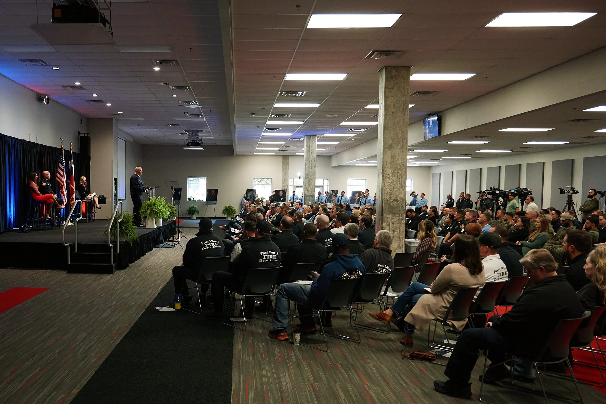 A large indoor gathering with a speaker at a podium on a stage, several people seated on the stage, and a diverse audience including fire department members in uniform and civilians, attending a formal event or ceremony.