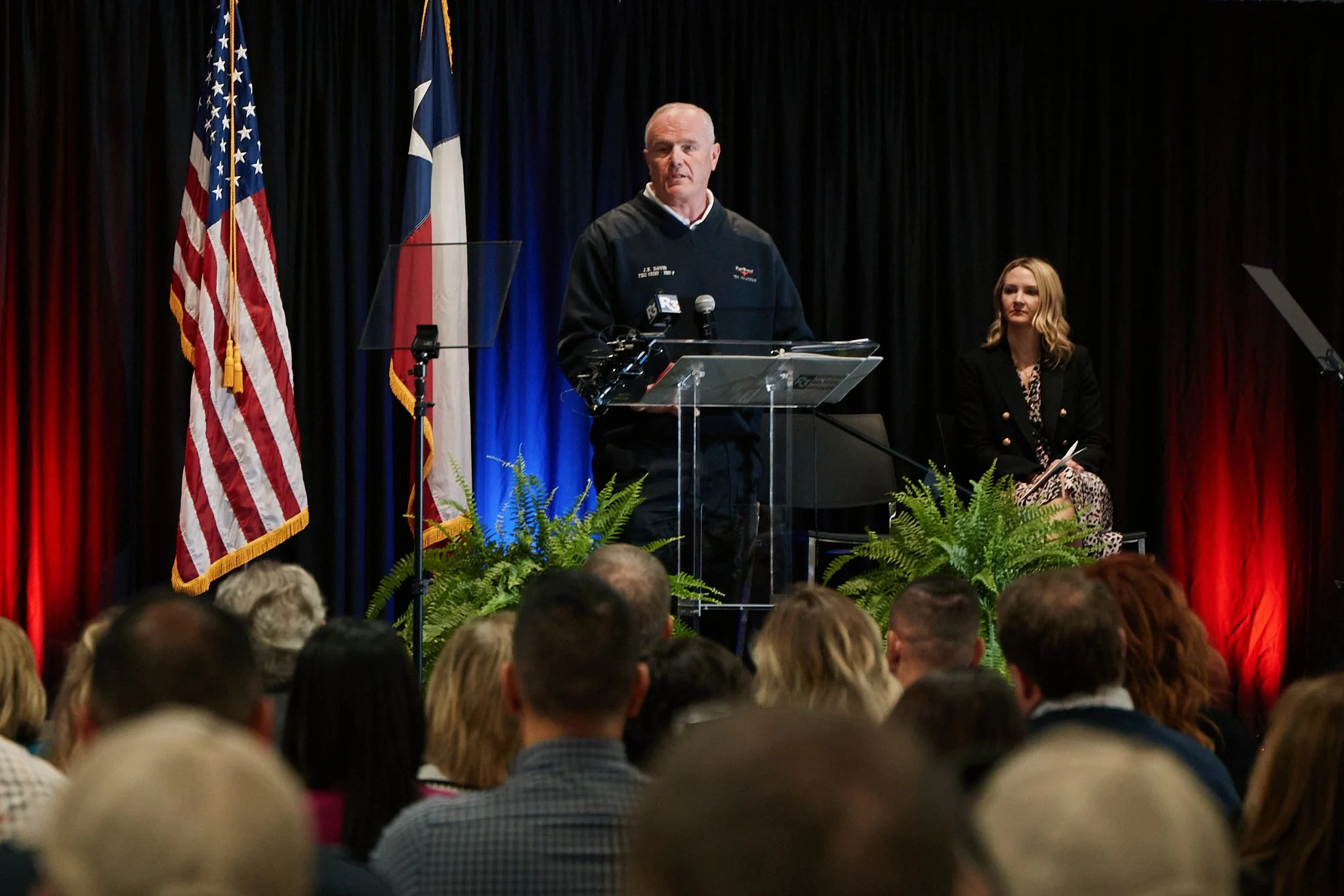 Man speaking at a podium on stage with black, red, and blue curtains, and flags of the United States and Texas behind him. Woman sitting to his right on stage.